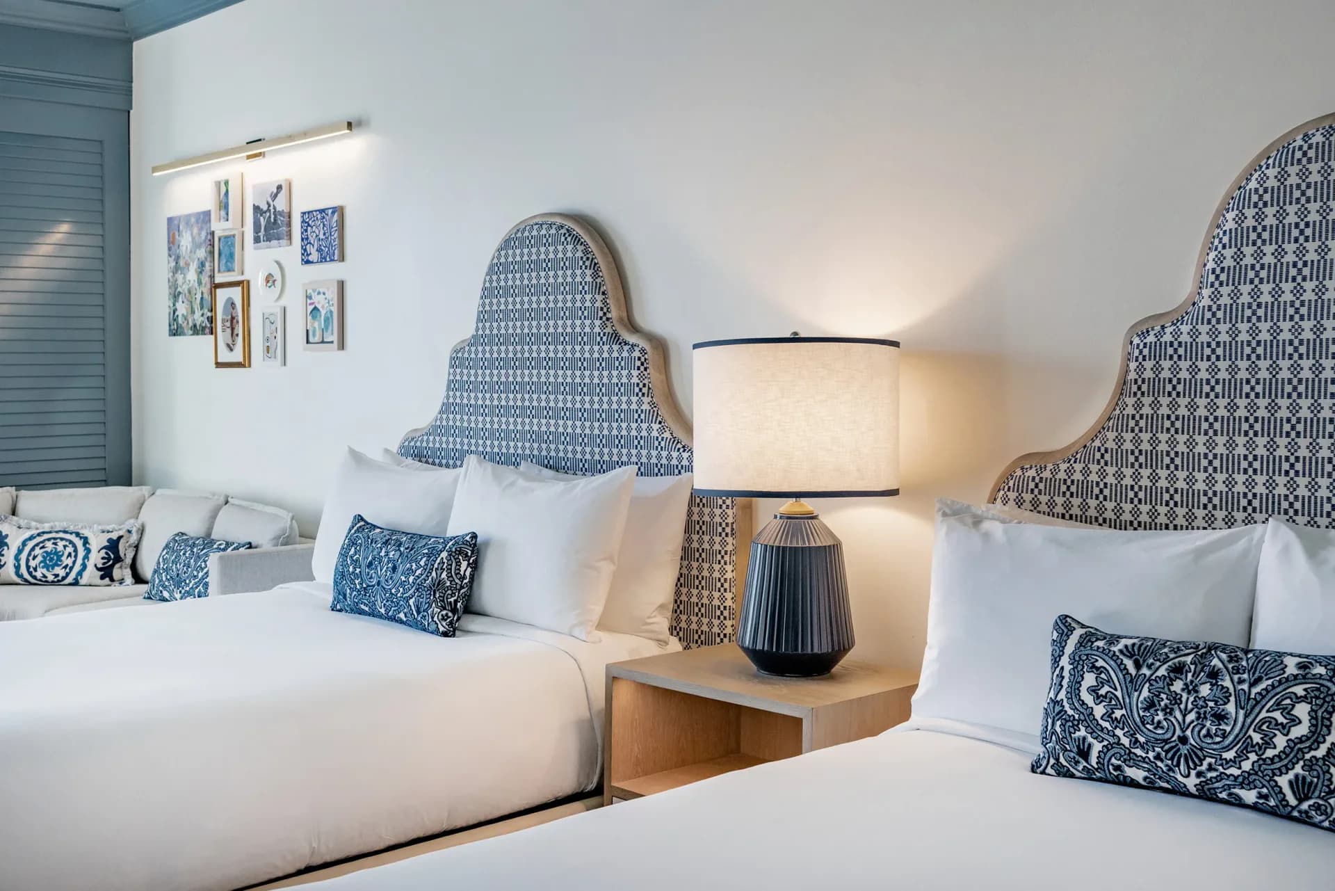 Modern hotel bedroom with two queen beds featuring blue patterned headboards, coordinating blue and white pillows, wood nightstands, and table lamps by Vanessa Watts.
