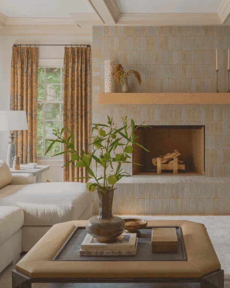 Neutral contemporary living room with striped wallcovering, wooden floating shelf, fireplace, golden drapery, and layered earth-tone palette by Victoria Bartholomew.