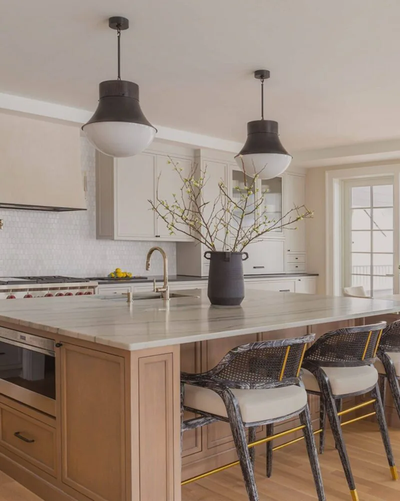 Modern kitchen with warm wood island, white countertops, dark pendant lights, and dining area with patterned chairs by Victoria Bartholomew.