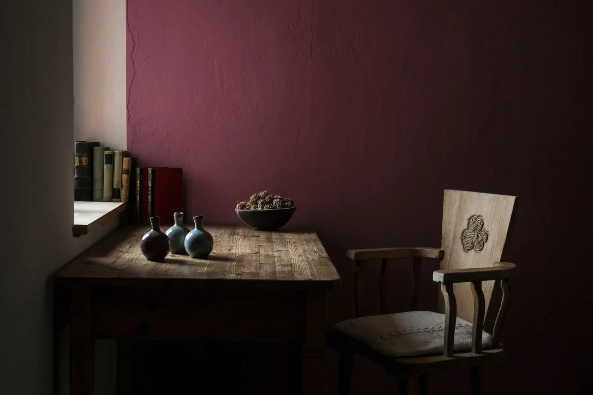 Moody interior scene with burgundy walls, wooden table displaying books and ceramics, chair with artwork, and dramatic lighting by Victoria Eremenko.