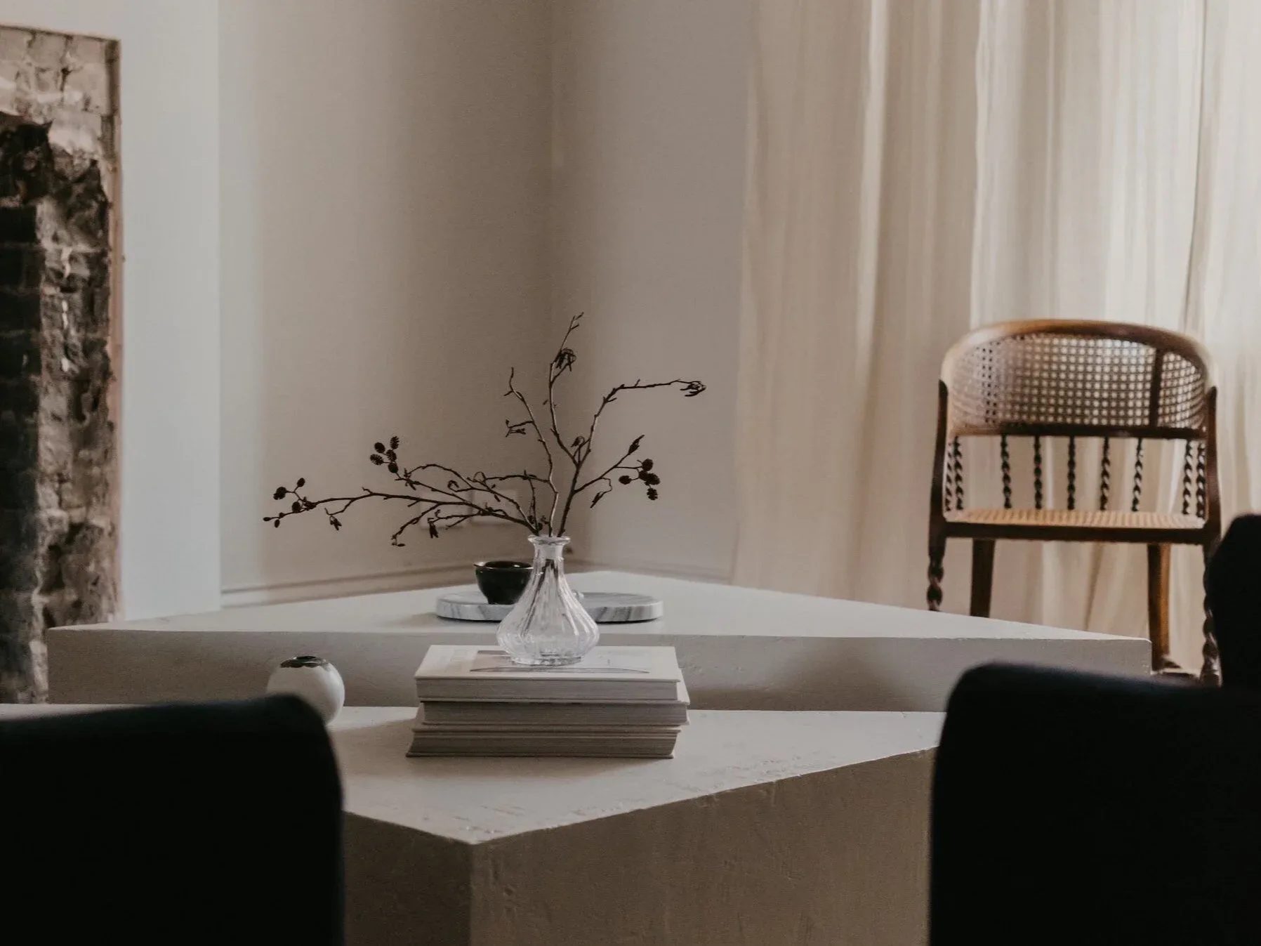 Minimalist interior with white cubic table, black chairs, rattan-back seating, dried branch arrangement in vase, and cream curtains by Victoria Eremenko.