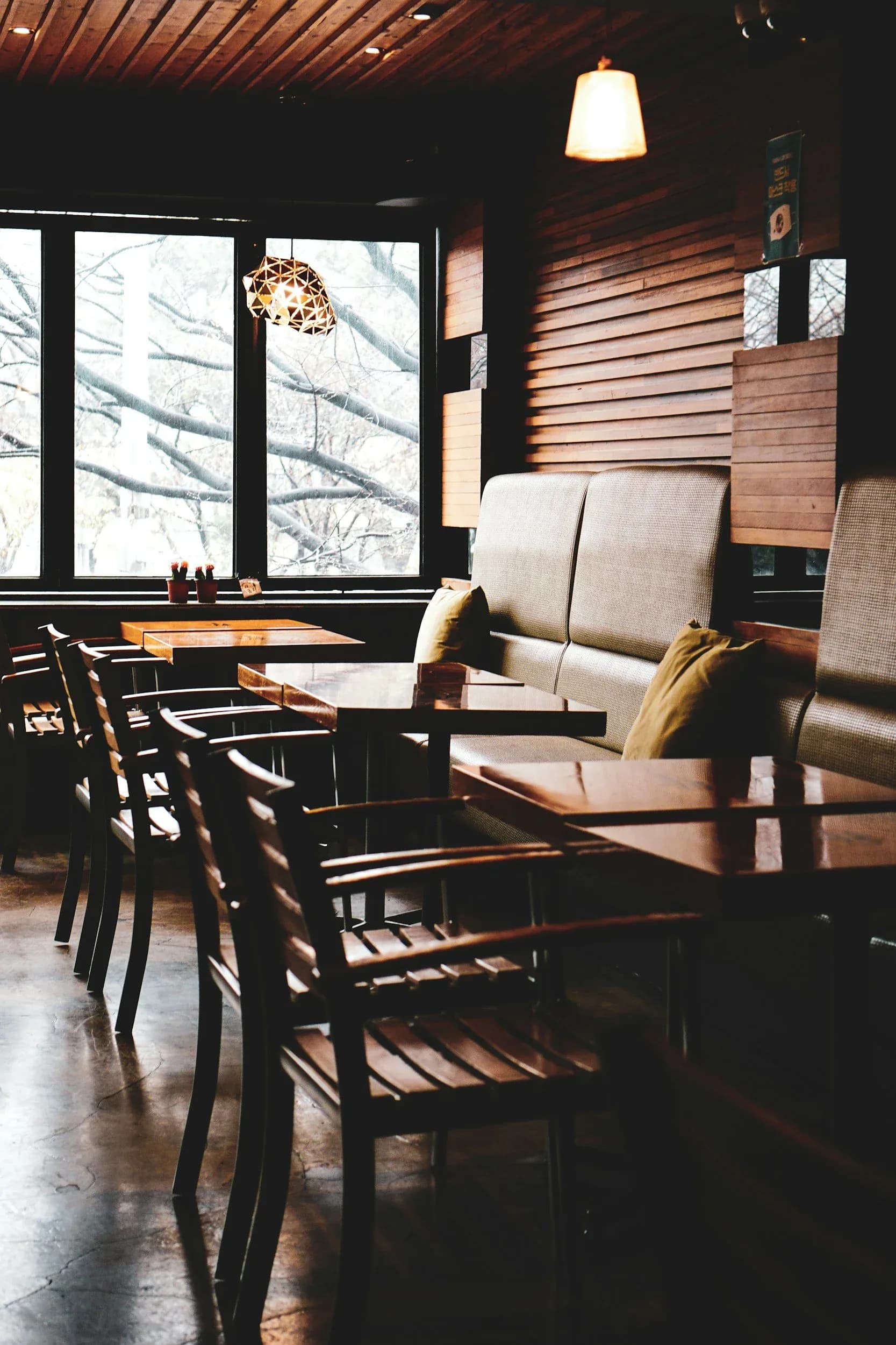 Modern dining area with black wooden chairs, beige upholstered banquette seating, wood plank walls, large windows overlooking winter landscape, and warm pendant lighting by Victoria Eremenko.