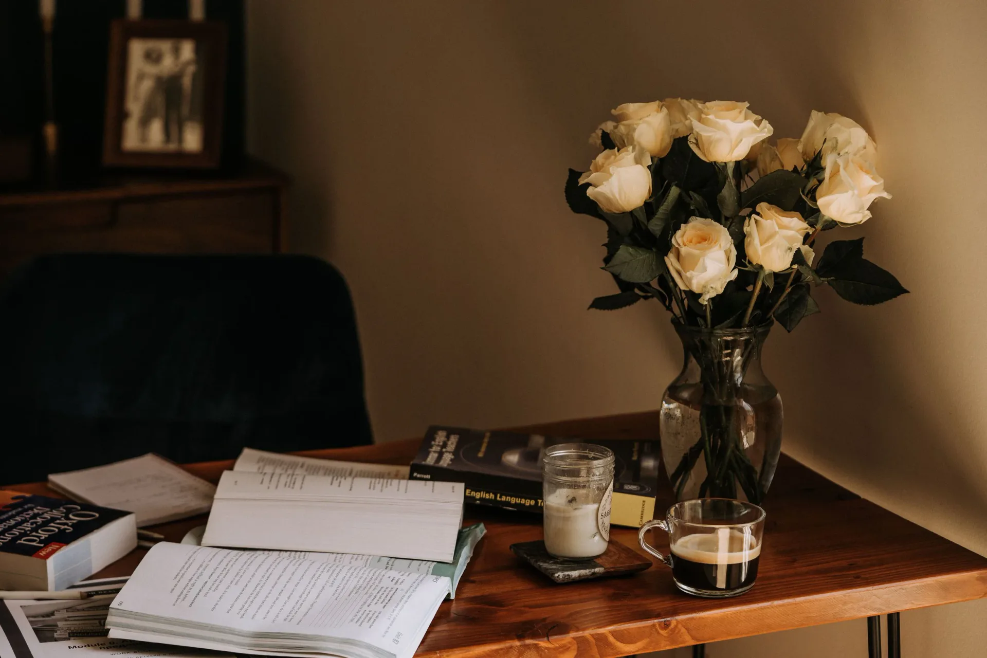 Warm-toned workspace with wooden desk featuring cream roses in glass vase, candles, and organized papers by Victoria Eremenko.
