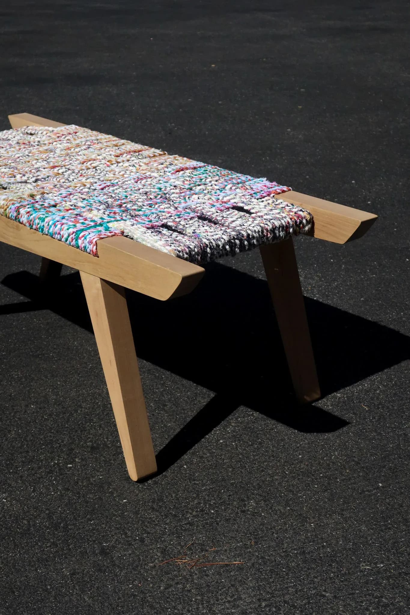 Light wood bench with woven textile seat in multicolored patterns, photographed outdoors on dark pavement by Victoria Humphrey.