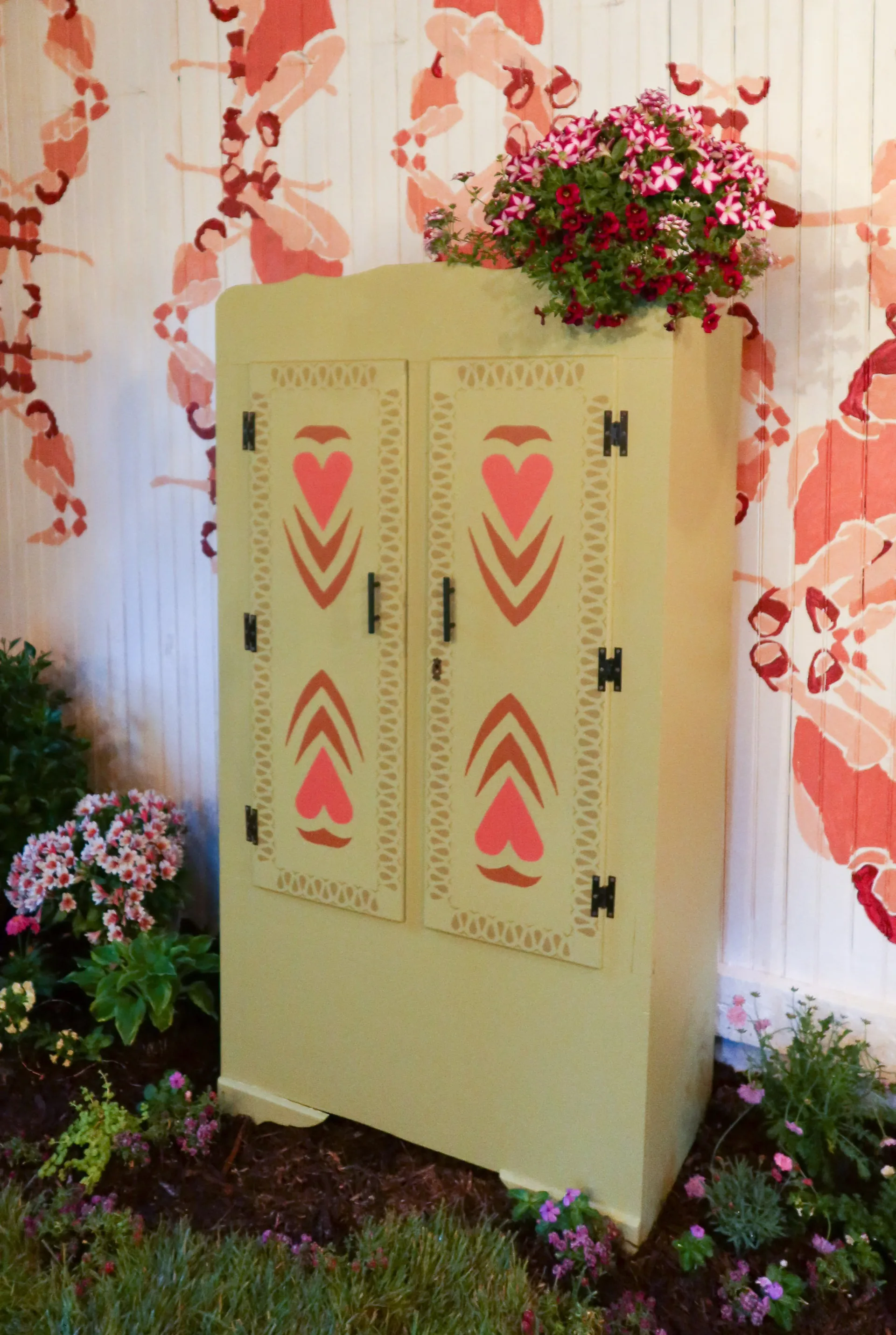 Pale yellow vintage refrigerator with red heart motifs and perforated detailing, surrounded by flowering plants against a patterned backdrop, by Victoria Humphrey.