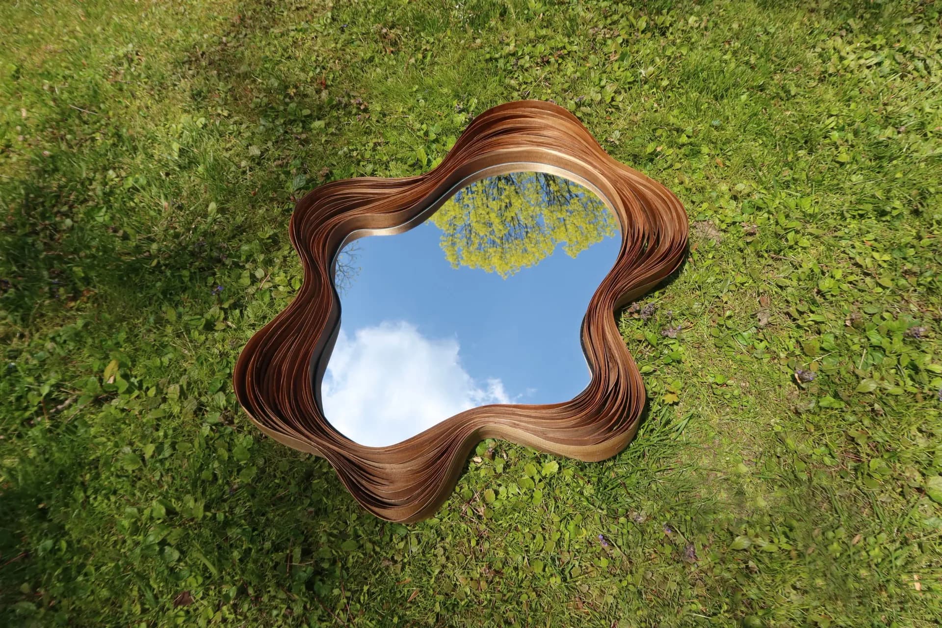 Ornate wooden wavy-edged mirror reflecting sky and clouds, installed on lush green moss-covered ground by Victoria Humphrey.