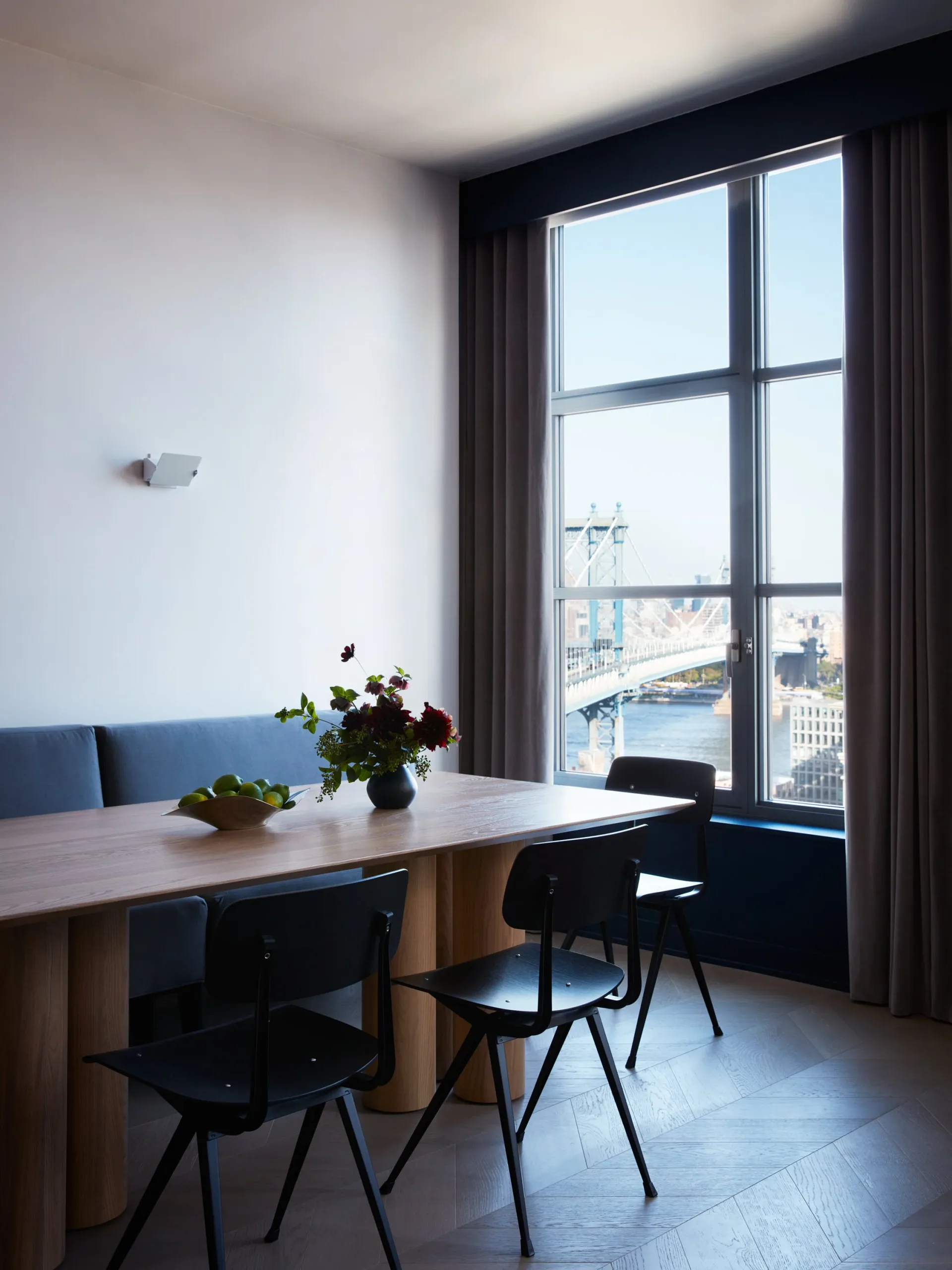 Modern dining space with wooden table, black chairs, and blue upholstered bench facing floor-to-ceiling windows overlooking a city waterfront, designed by Vinh Nguyen.