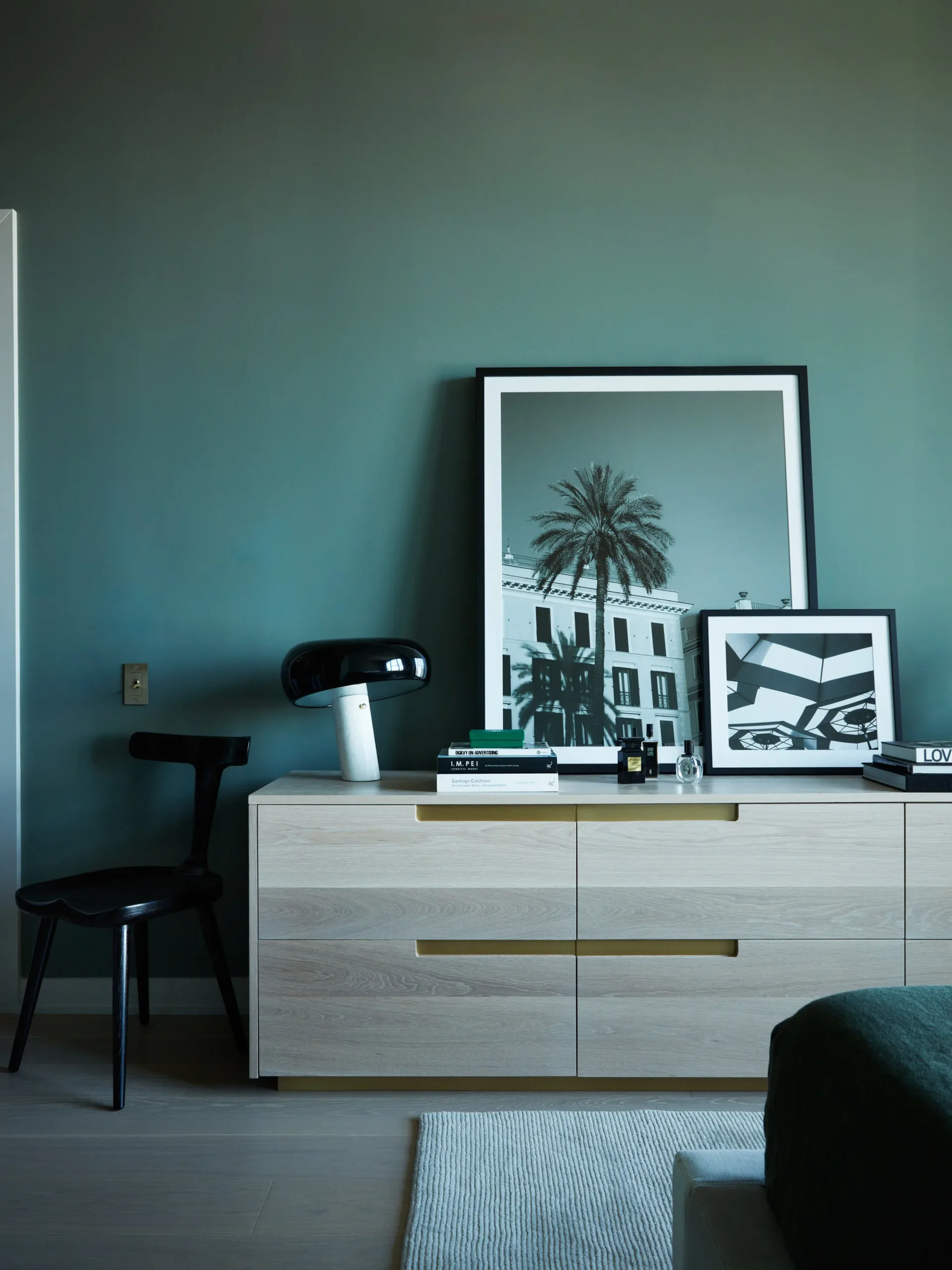 Modern bedroom interior with turquoise walls, white dresser with brass hardware, black chair, framed palm tree photograph, and minimal black accessories by Vinh Nguyen.