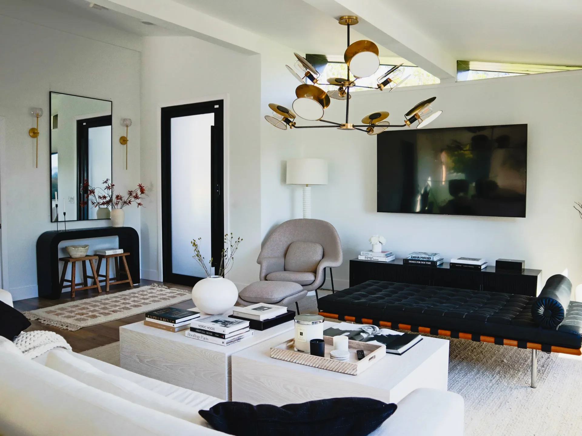 Modern living room with white walls, black media console, curved grey armchair, brass chandelier, and black-framed mirrors by Vinh Nguyen.