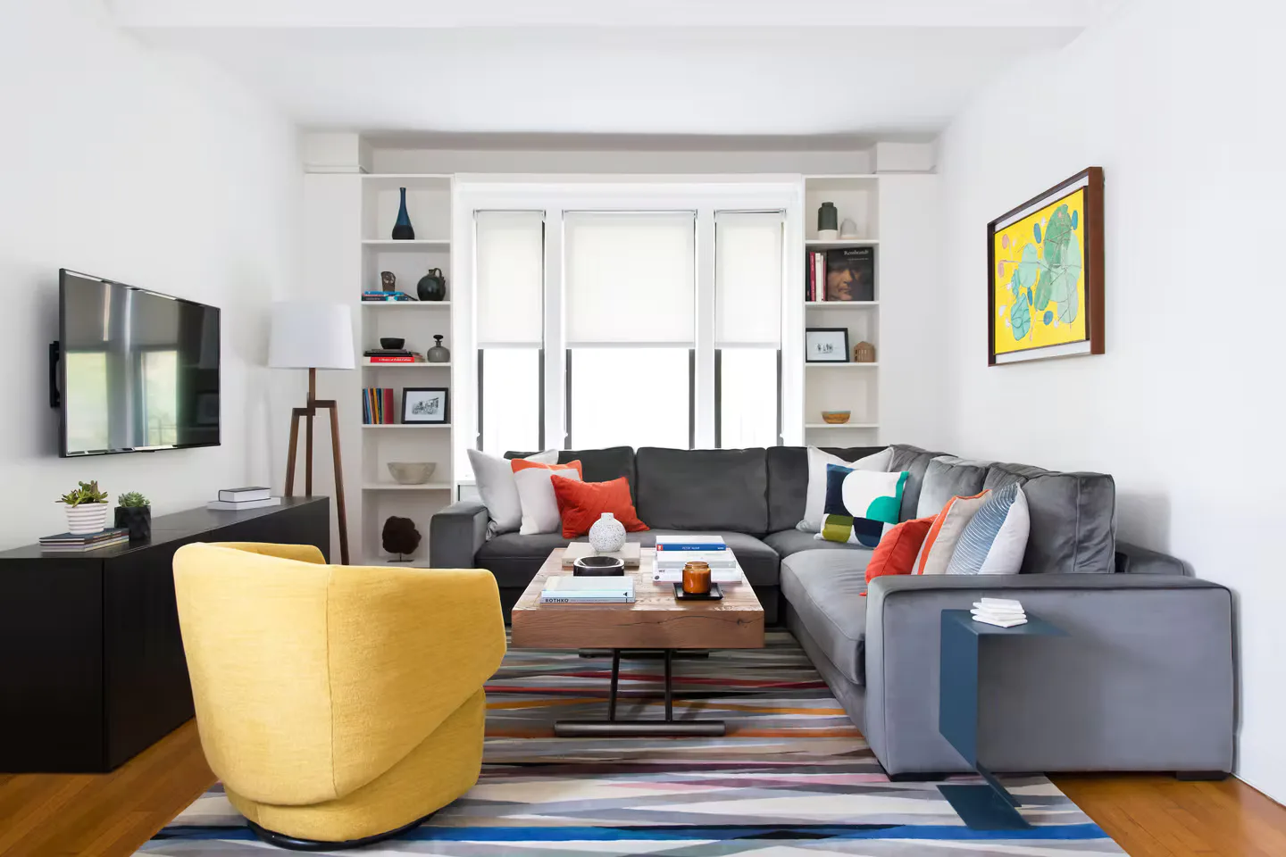 Modern living room with gray sectional, yellow curved chair, wood floors, and built-in shelving by Vivian Bayles.