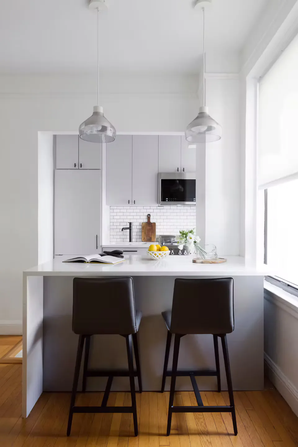 Modern white kitchen with marble countertop, subway tile backsplash, pendant lighting, and black bar stools by Vivian Bayles.