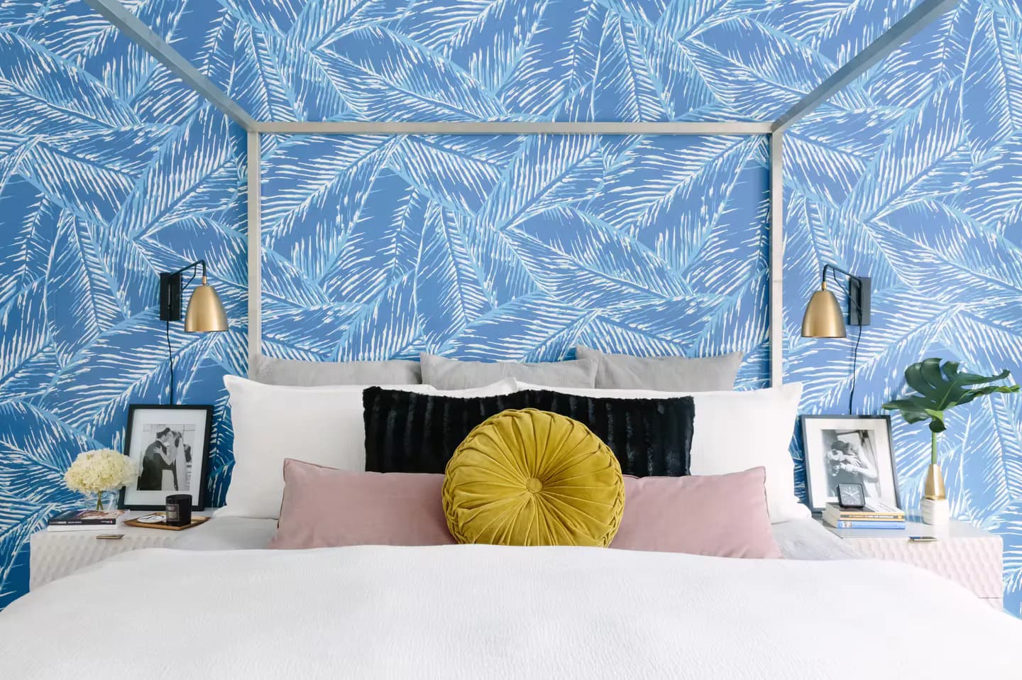 Modern bedroom with blue palm-leaf wallpaper, white canopy bed, black headboard, pink and white bedding, and yellow accent pillow by Vivian Bayles.