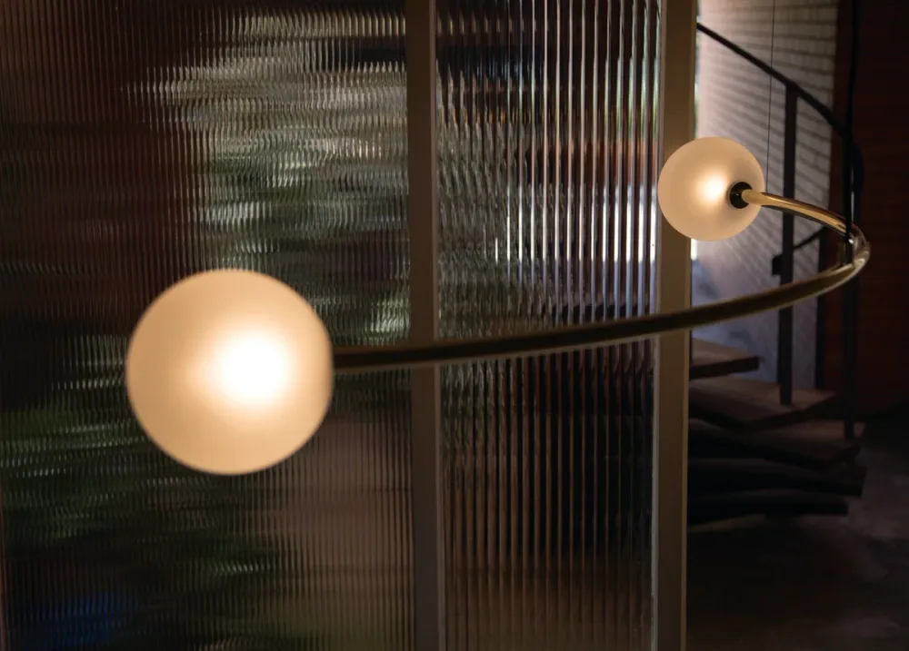 Modern pendant lighting installation with frosted glass spheres suspended from curved brass arms against textured wall panels by Volker Haug.