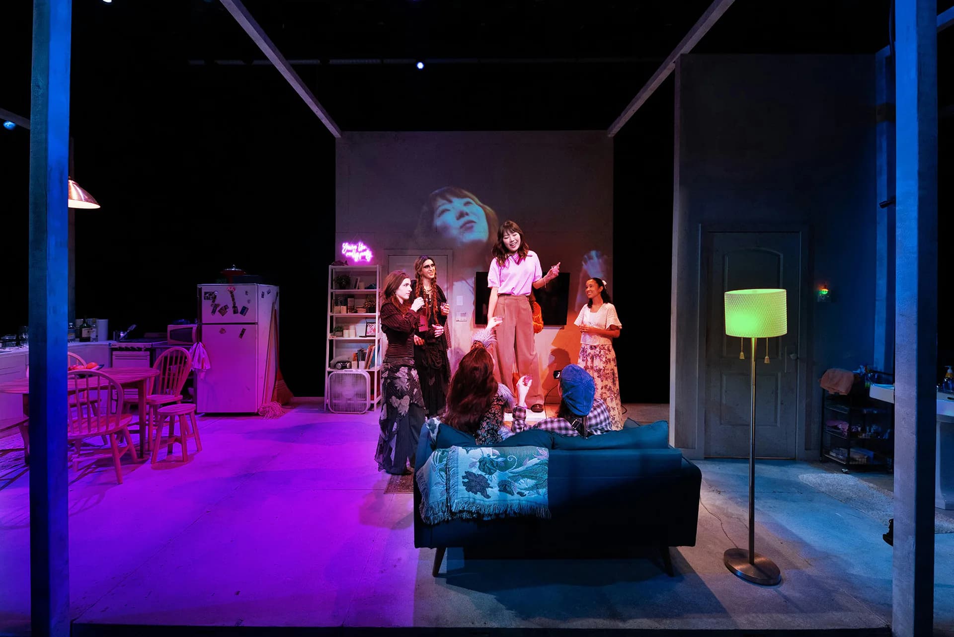 A theatrical stage set with vibrant purple and blue lighting, featuring furniture, shelving, and costumed figures in a contemporary interior design by Weniz Wills.