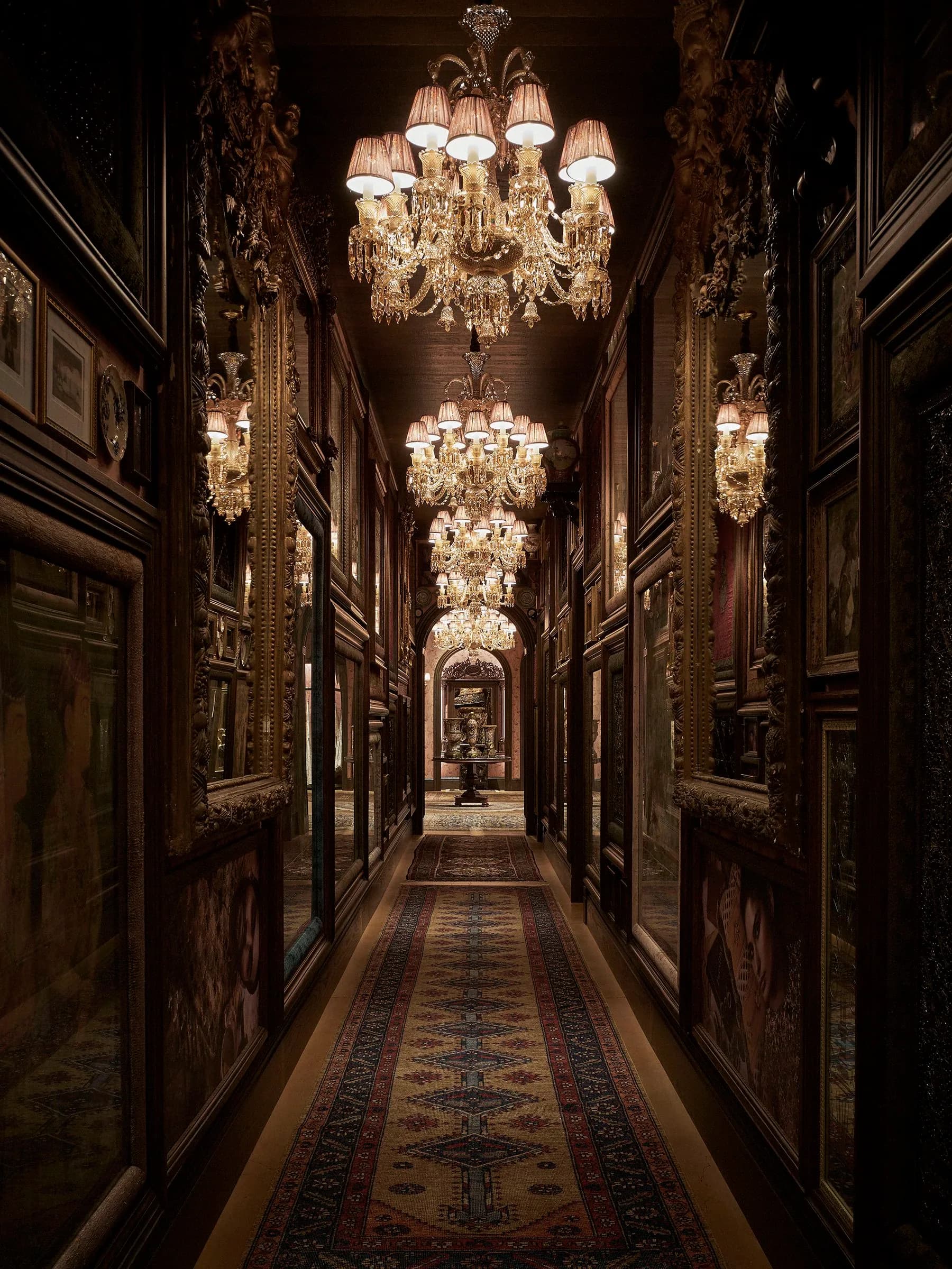 A luxurious hallway with ornate dark wood paneling, crystal chandeliers, and a patterned runner rug stretching into the distance by William Armstrong.