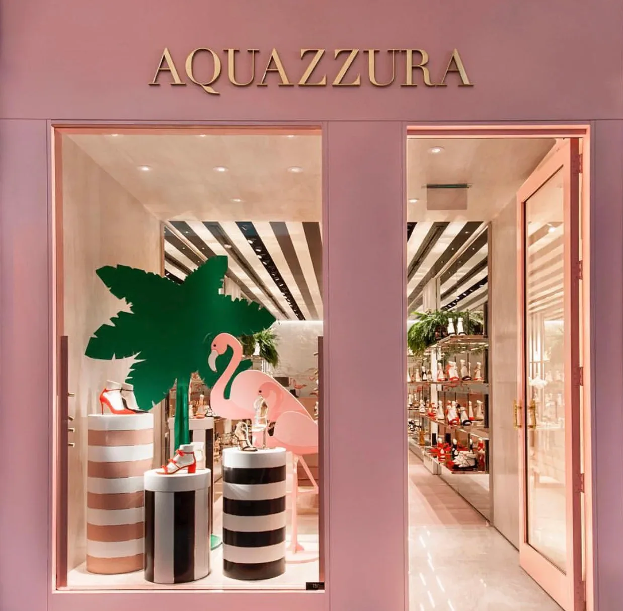 Aquazzura retail storefront with pink façade, striped interior walls, green palm tree installation, and striped cylindrical display pedestals by William Armstrong.