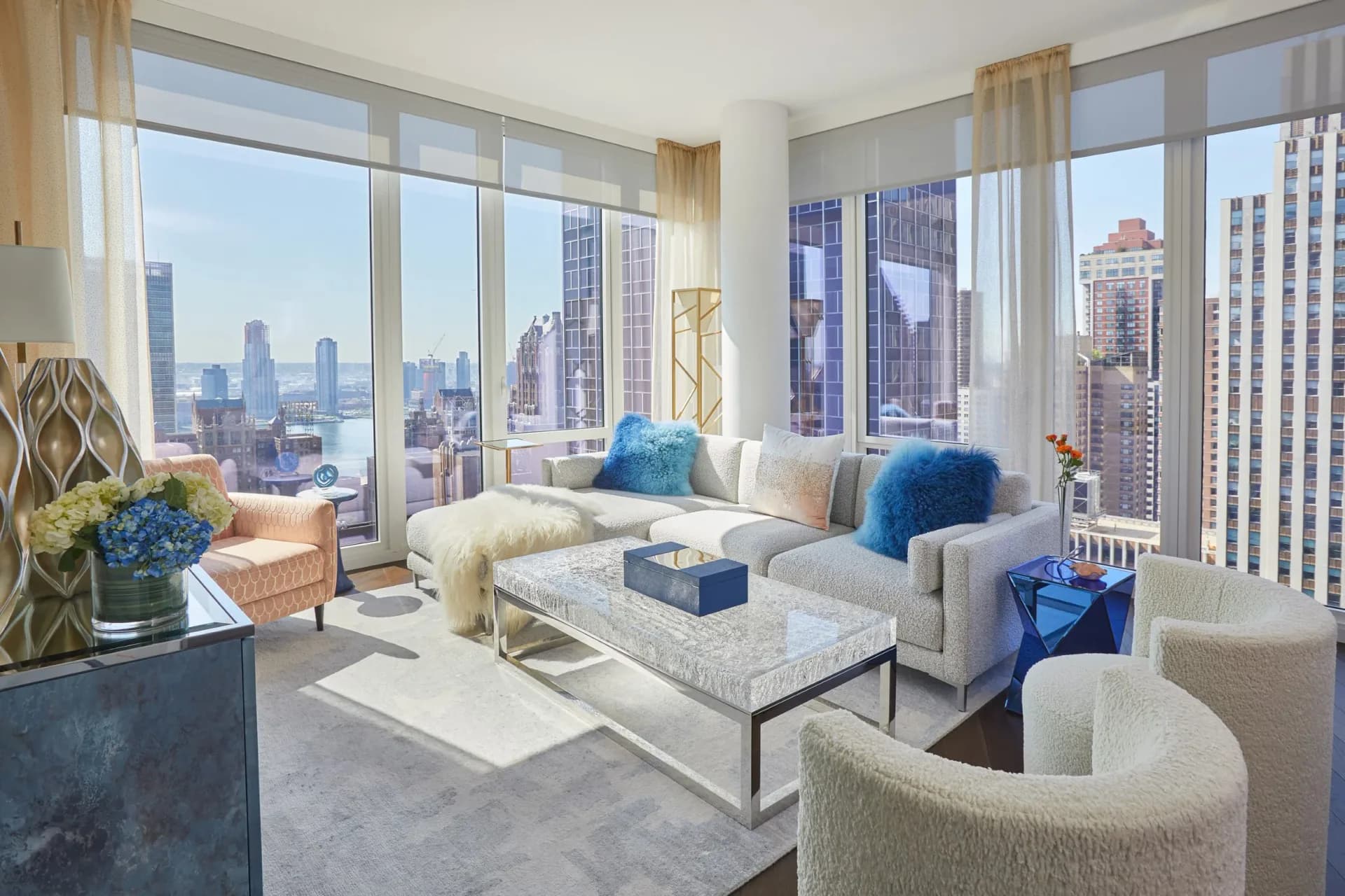 High-rise residential living room with floor-to-ceiling windows overlooking a city skyline, featuring cream and blue upholstered furniture, marble accents, and warm lighting by Yasemin Turckan.
