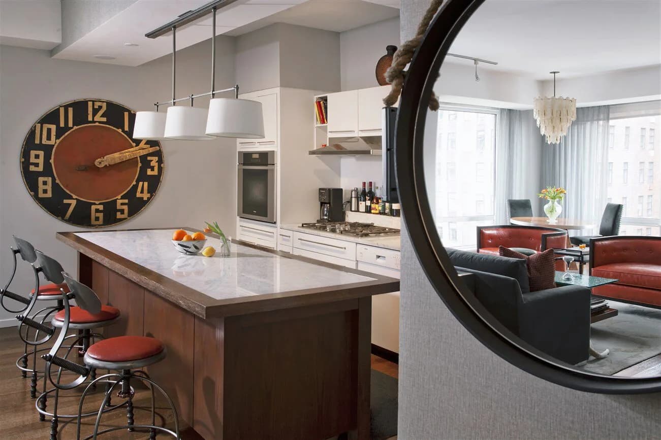 Modern kitchen and living area with dark wood island, white cabinetry, geometric wall clock, large round mirror, and burgundy seating by Yasemin Turckan.