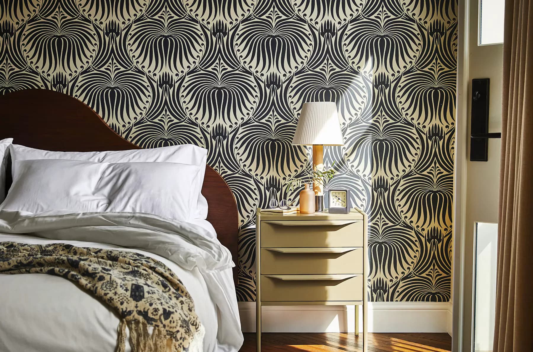 Modern bedroom with bold black and gold geometric patterned wallpaper, white bedding, wooden nightstand with table lamp, and hardwood flooring by Yasmin Murtada.