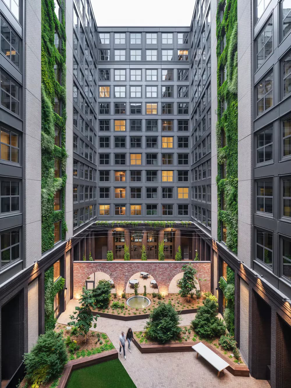 Courtyard of a modern residential building with brick paving, green planters, lounge seating, and climbing ivy on surrounding concrete facades by Yasmin Murtada.