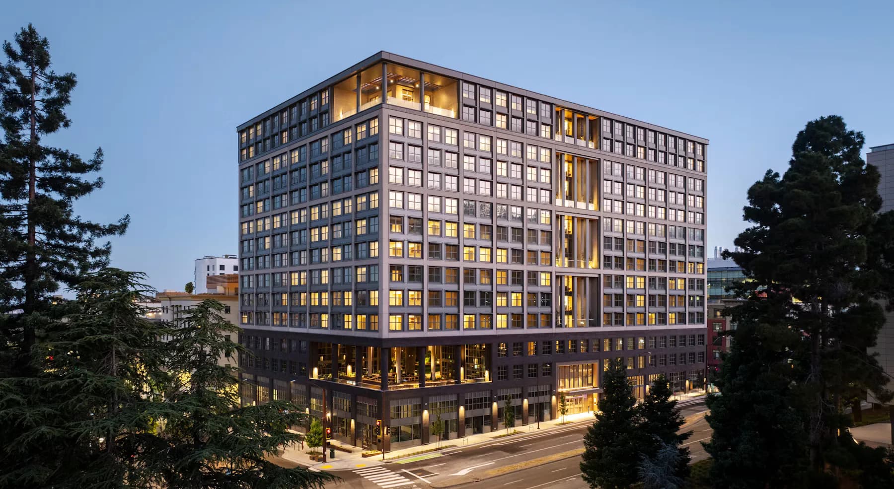 Modern multi-story residential building with golden-toned facade and illuminated ground-floor retail, surrounded by trees at dusk, by Yasmin Murtada.