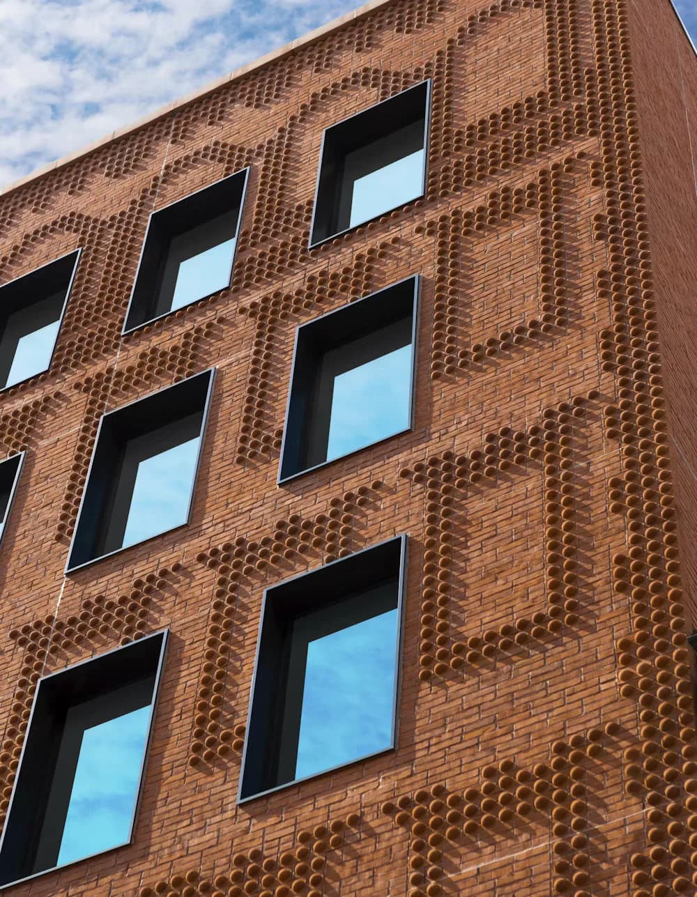 Brick facade with geometric perforated pattern and recessed windows reflecting sky, showcasing contemporary architectural detailing by Yasmin Murtada.