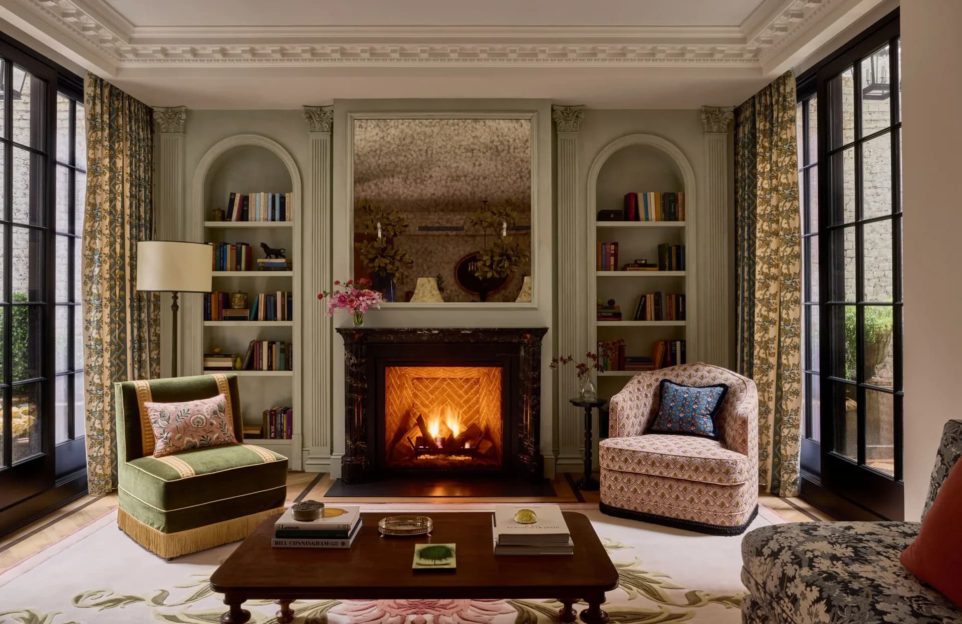 Elegant living room with symmetrical built-in shelving, fireplace with glowing fire, patterned armchairs, and tall windows with gold curtains by Young Huh.