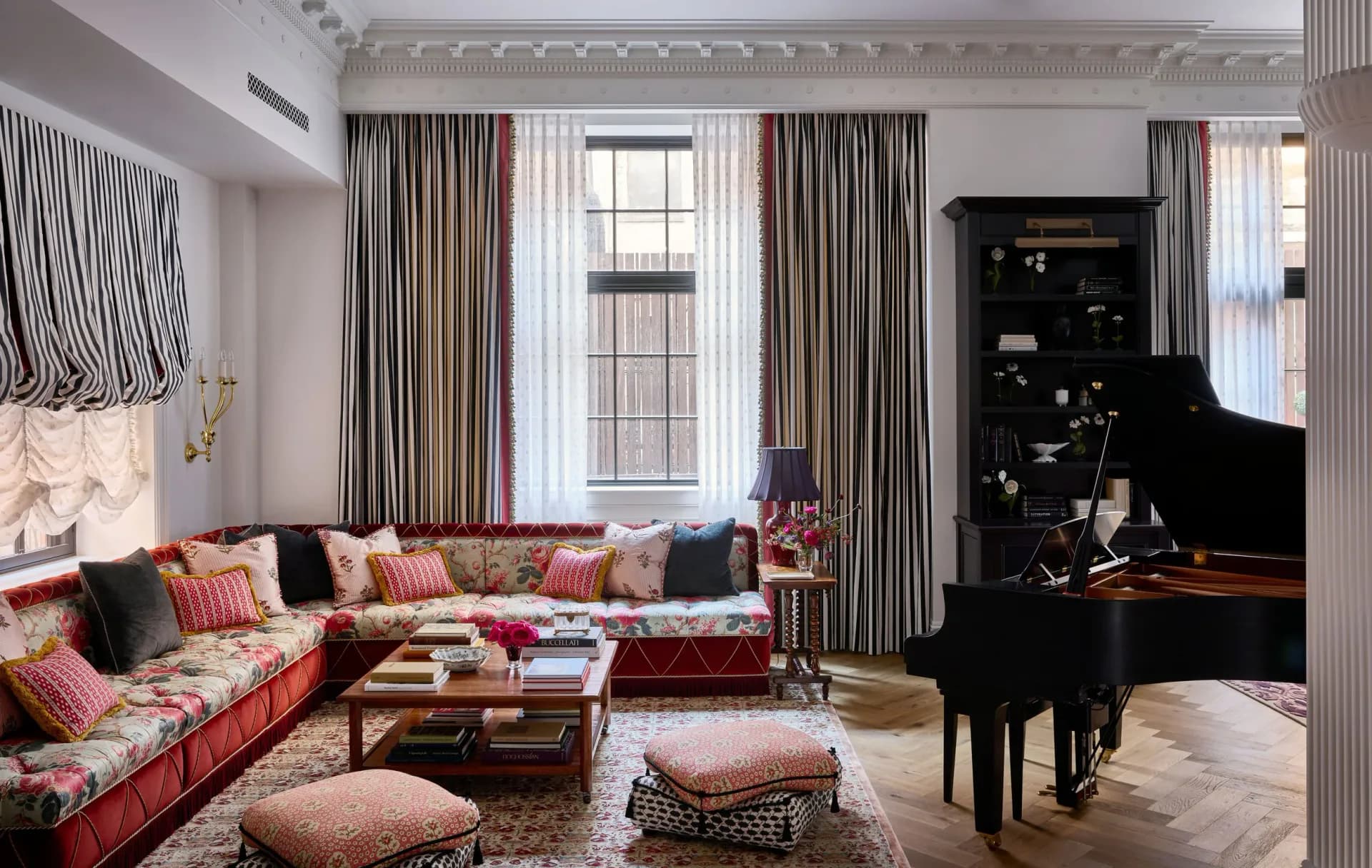 Luxurious living room with coral-red sectional, striped curtains, black grand piano, and patterned ottomans by Young Huh.