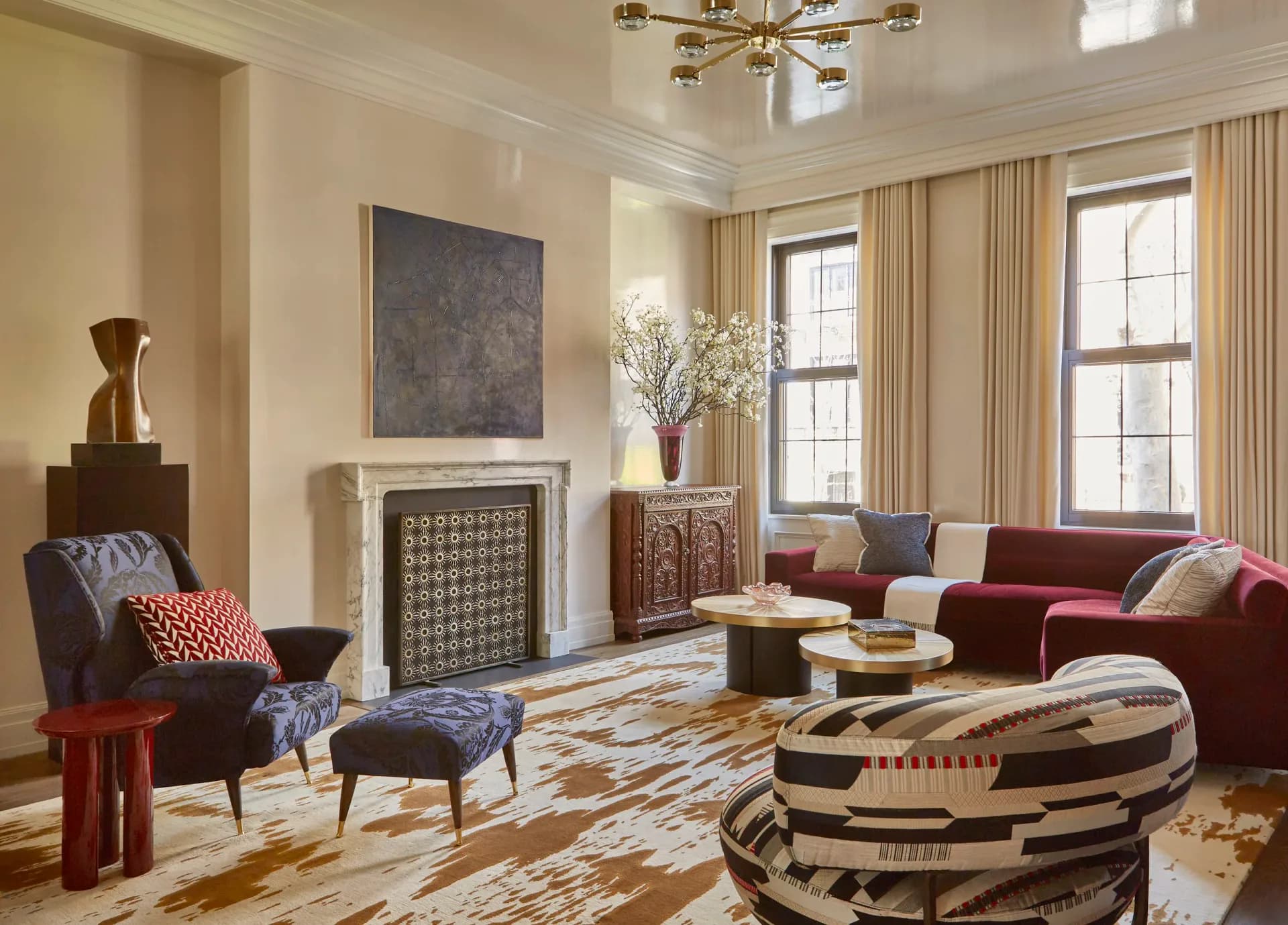 Sophisticated living room with burgundy sofa, patterned rug, fireplace, and cream curtains in warm neutral palette by Young Huh.