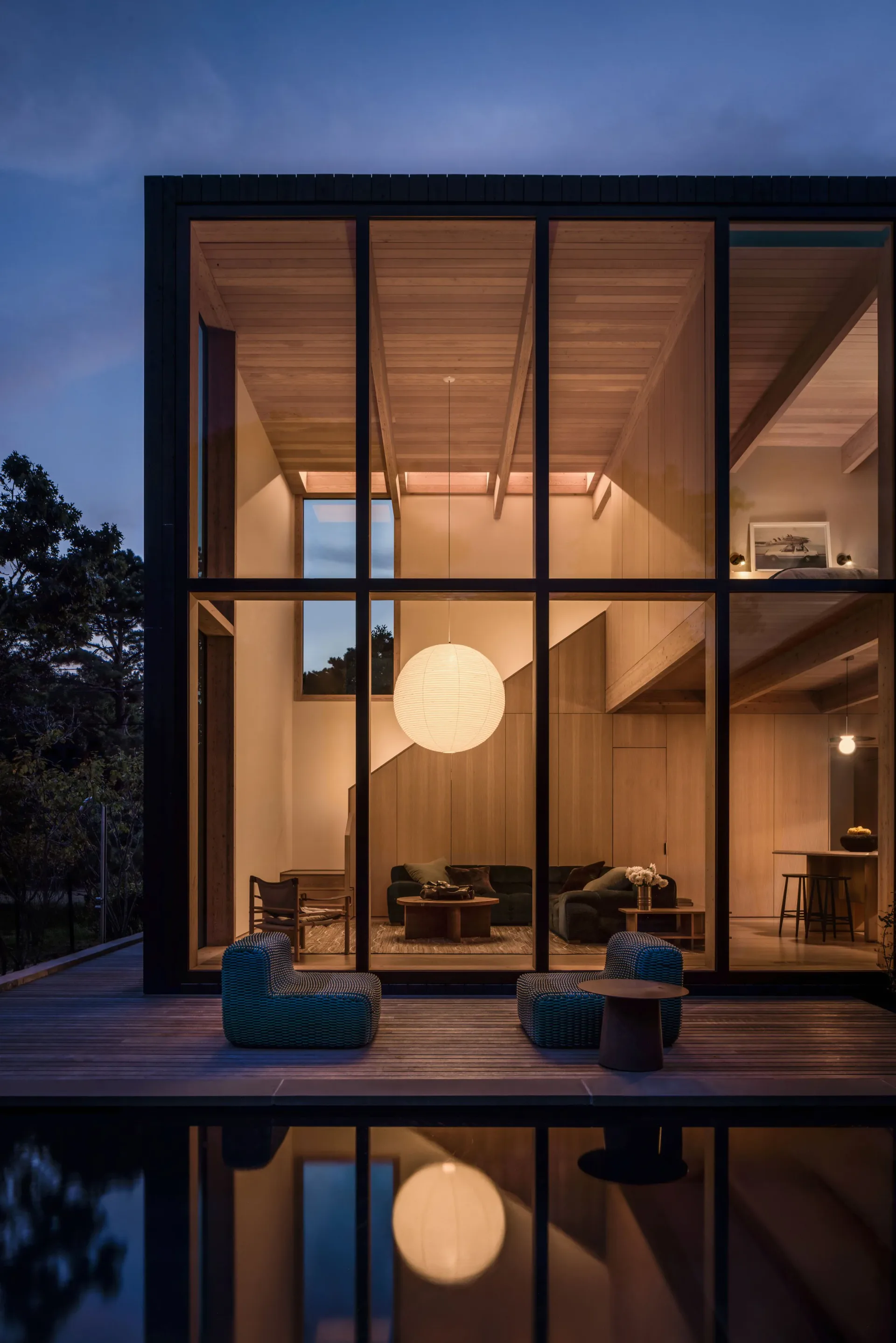 Modern minimalist house with black-framed glass walls, warm wood interior lighting, and reflecting pool at dusk by Yubi Park.