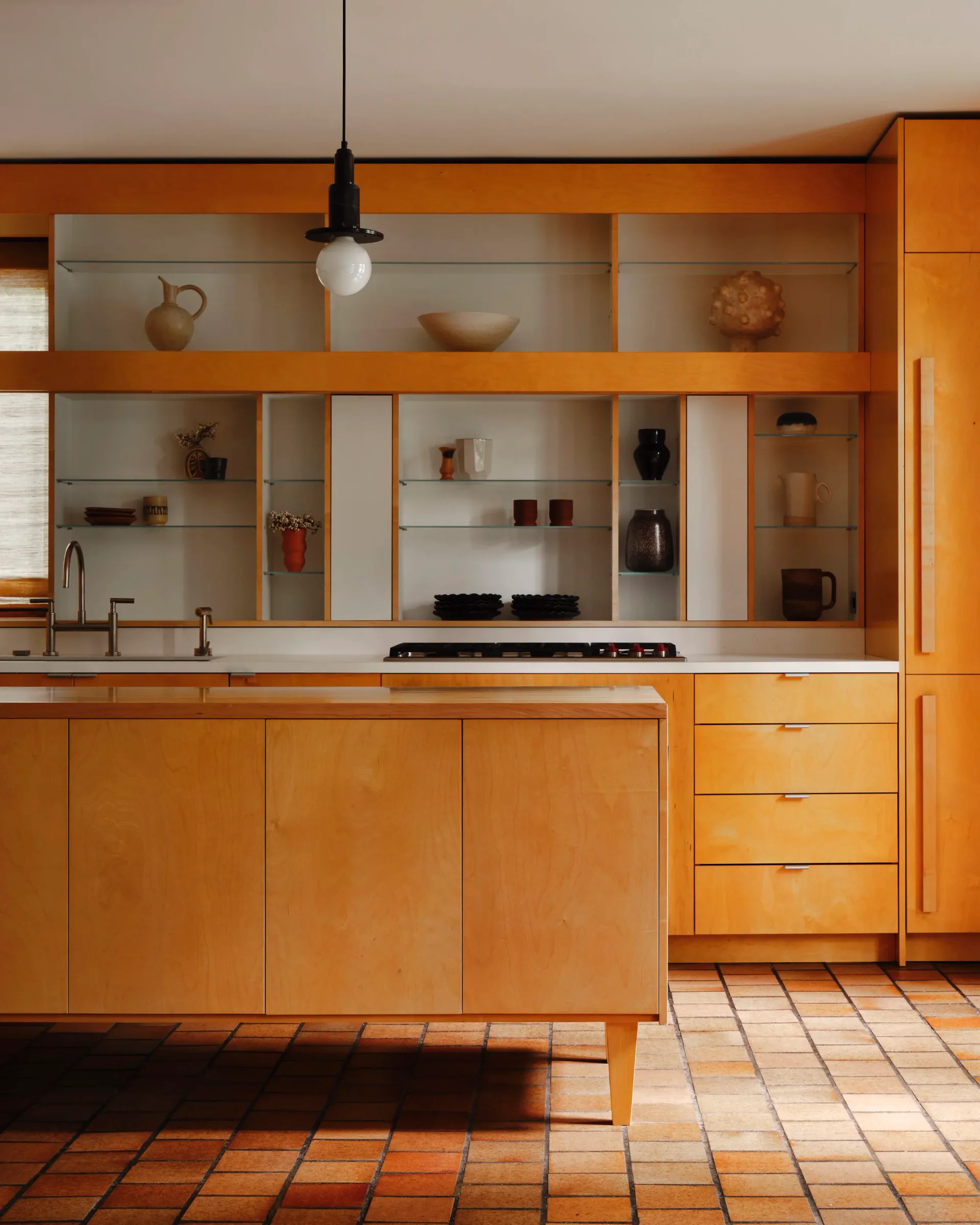 Warm-toned modern kitchen with natural wood cabinetry, open shelving, terracotta tile flooring, and minimalist pendant lighting by Yubi Park.