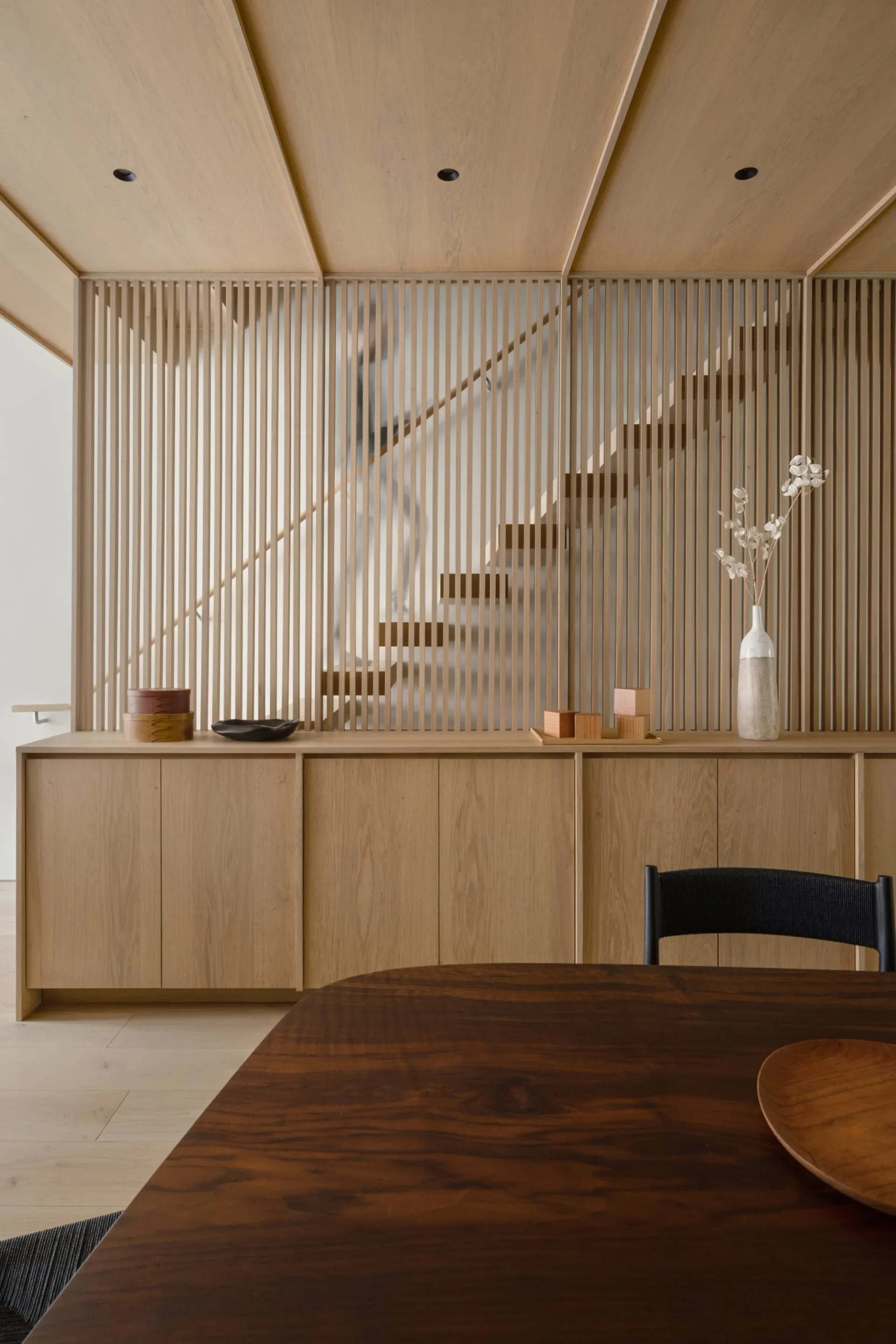 Minimalist interior with light wood cabinetry, vertical slatted wall panels, floating staircase, and dining table in warm neutral tones by Yubi Park.