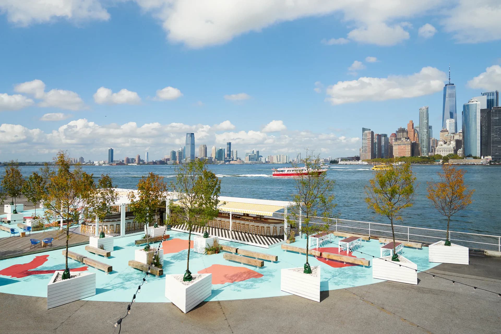 Waterfront plaza with light blue paving, geometric seating elements, and young trees overlooking Manhattan skyline and Hudson River, designed by Yubi Park.