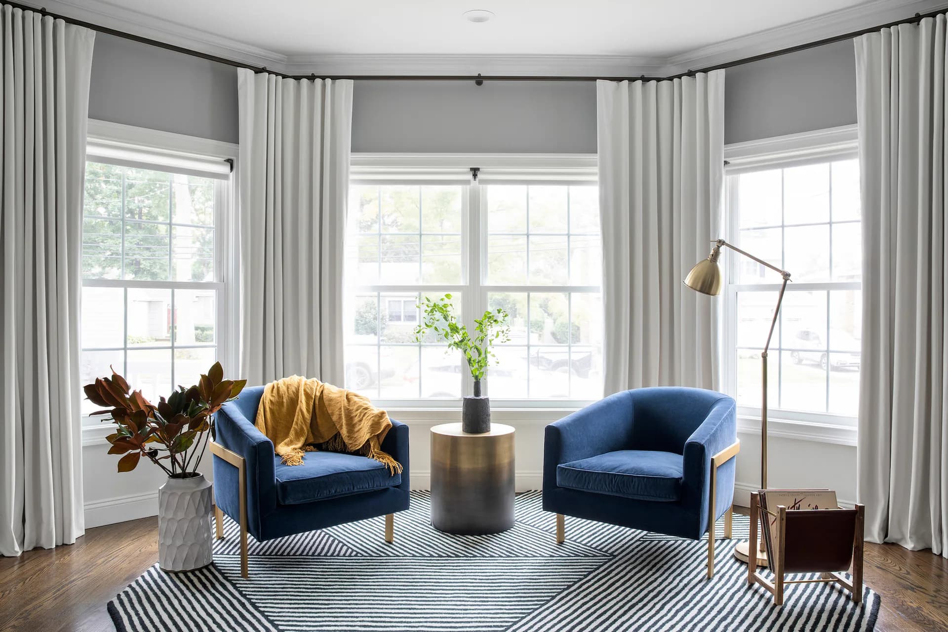 Bright bay-window seating area with two navy blue velvet armchairs, brass side table, striped area rug, and floor lamp, featuring floor-to-ceiling curtains and multiple windows by Yunchi Jong.