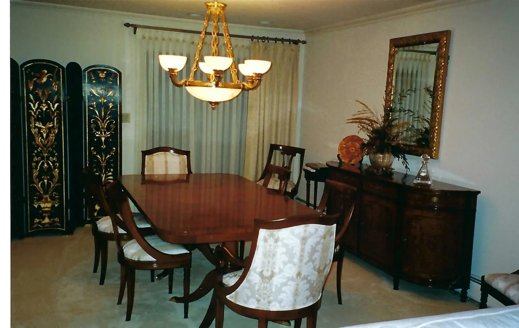 Formal dining room with mahogany table, upholstered chairs, brass chandelier, decorative wall panels, and dark wood sideboard by Yvonne Compitello.