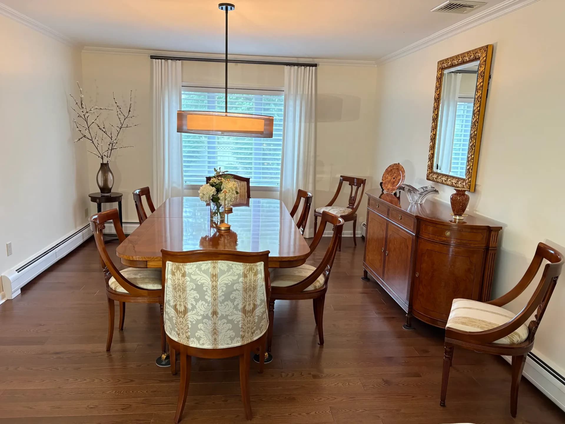 Warm dining room with round upholstered table, wooden chairs, dark wood sideboard, and glass doors overlooking water, designed by Yvonne Compitello.