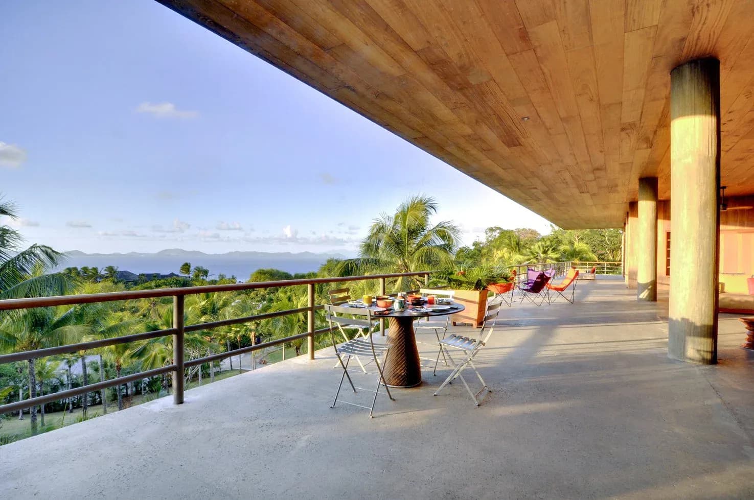 Expansive modern concrete terrace with metal railings overlooking lush tropical landscape and distant mountains, featuring casual seating areas beneath a striking wooden soffit by Zachary Lewis.