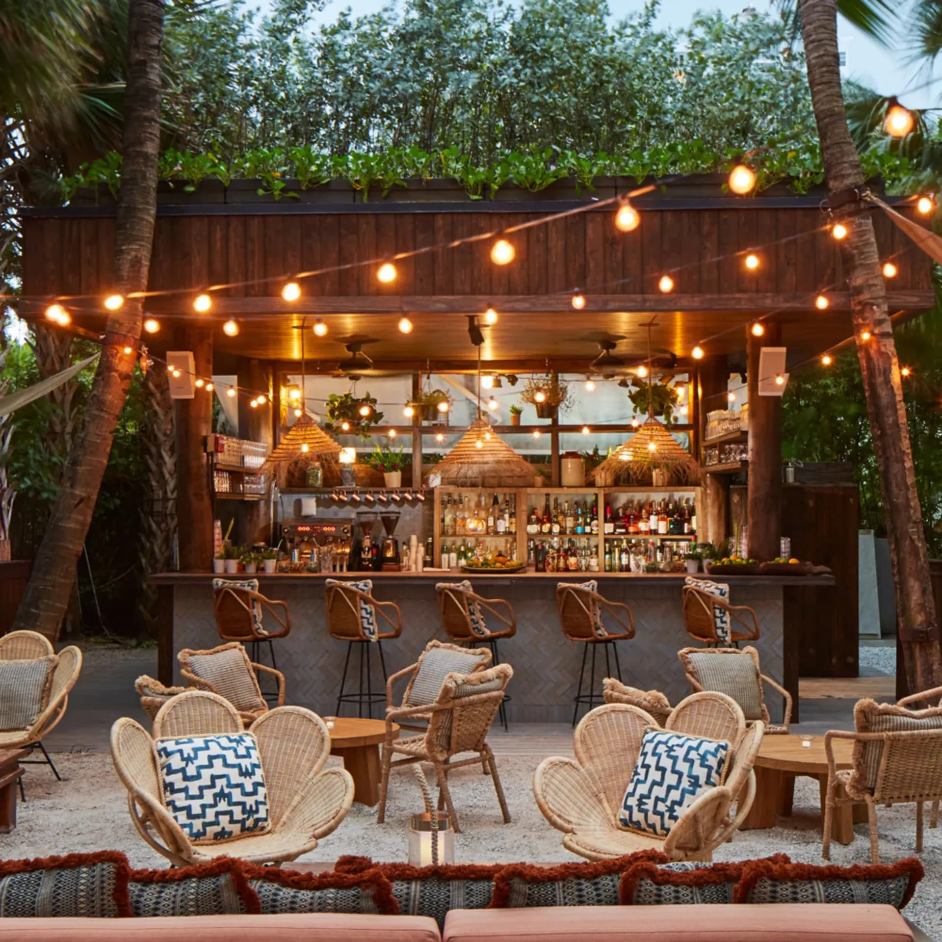 Tropical outdoor bar and lounge with wooden pergola, string lights, rattan seating, and palm trees by Zak Profera.