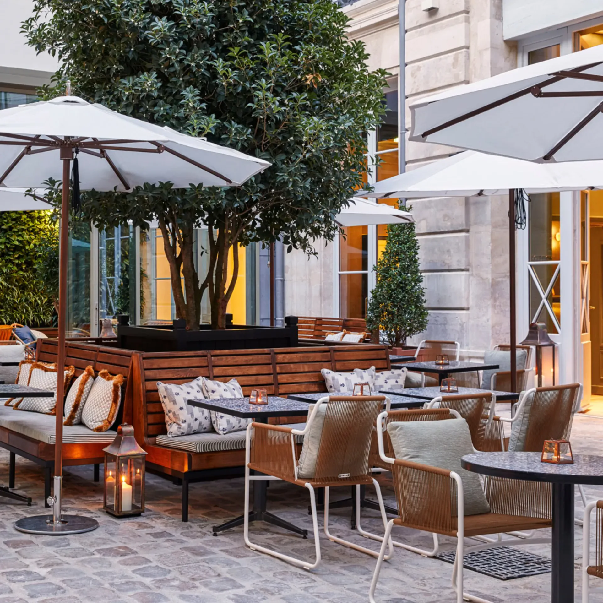 Elegant outdoor dining terrace with wooden built-in banquette seating, metal chairs, black tables, umbrellas, and mature tree canopy by Zak Profera.
