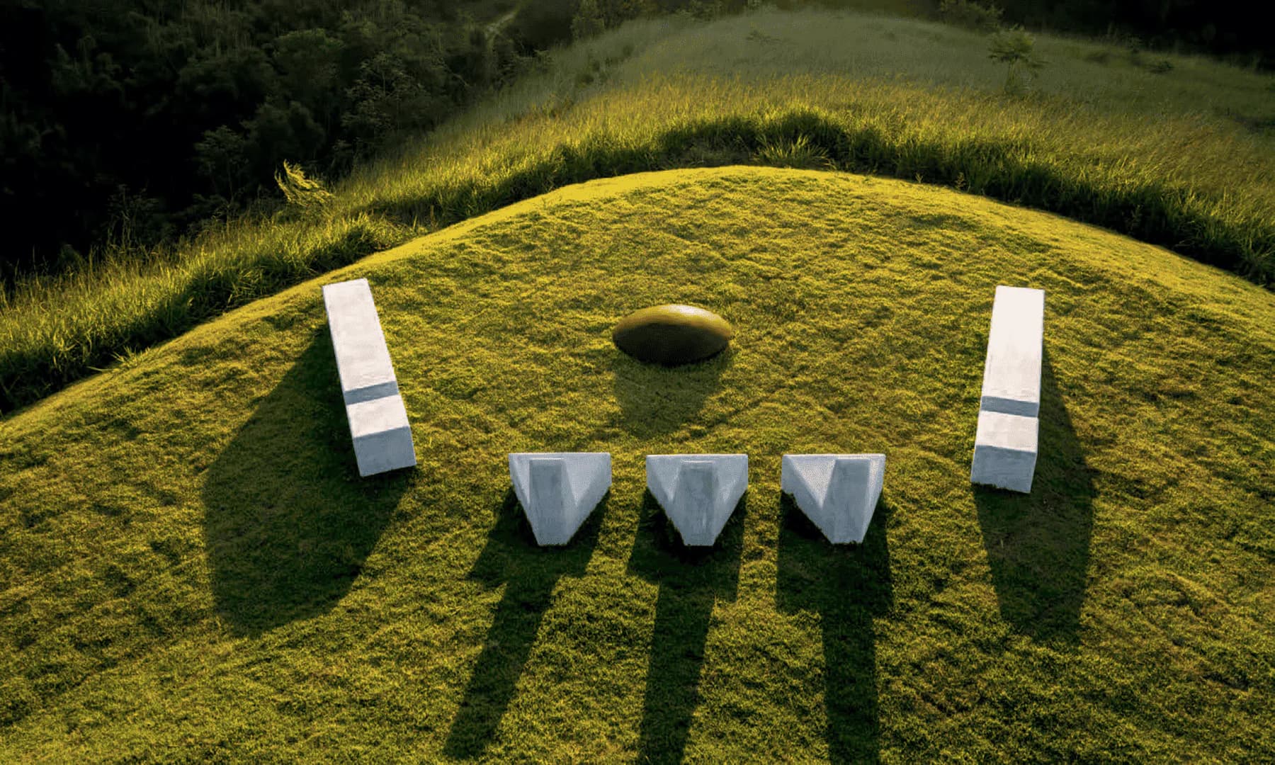 Sculptural landscape installation with white geometric forms arranged on moss-covered earthwork mound, creating striking shadows across verdant hillside by Zanini de Zanine Caldas.