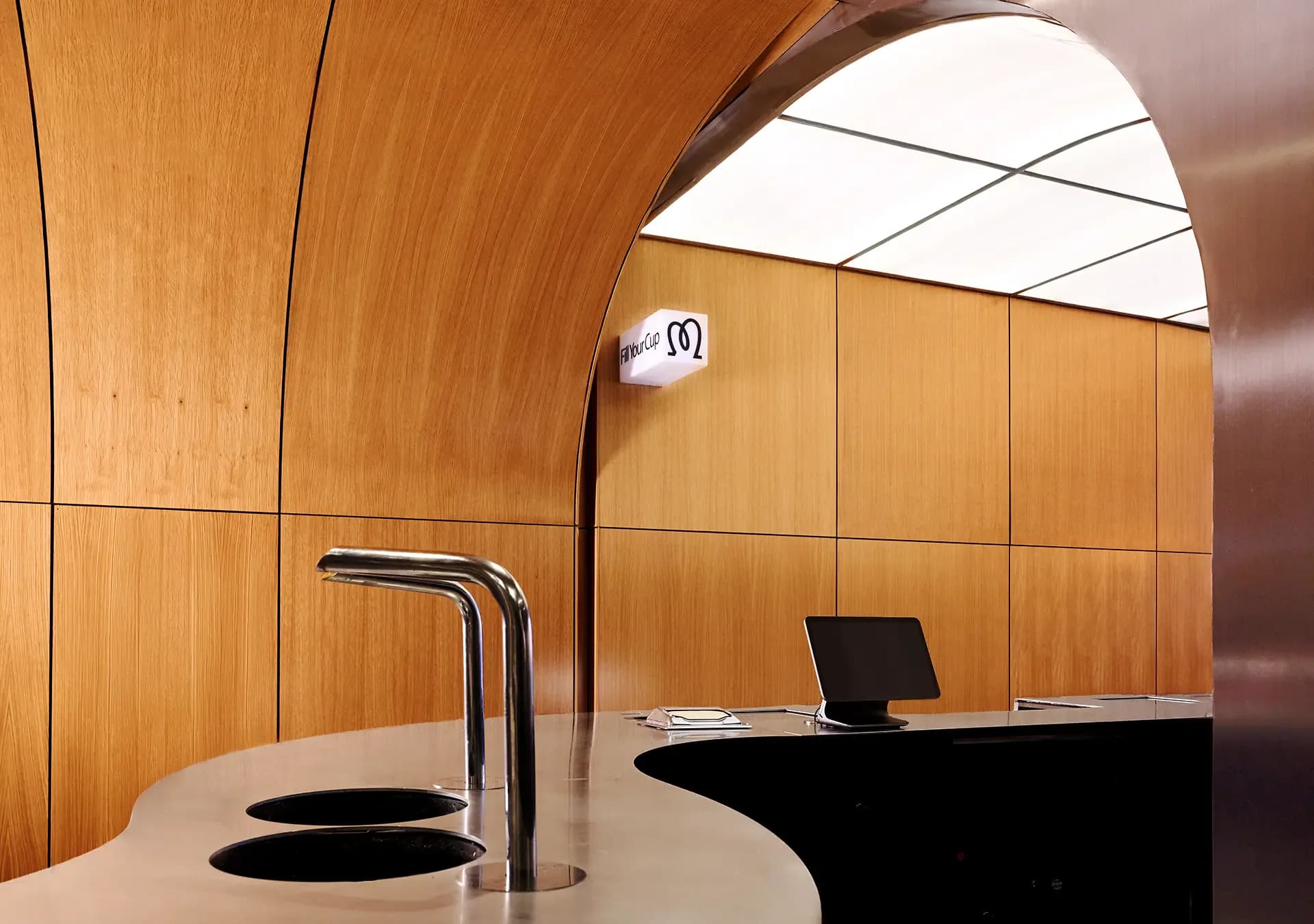 Modern curved wooden interior with arched ceiling, illuminated skylight, curved counter with black circular elements, chrome faucet, and laptop workspace by Zhixiao Yu.