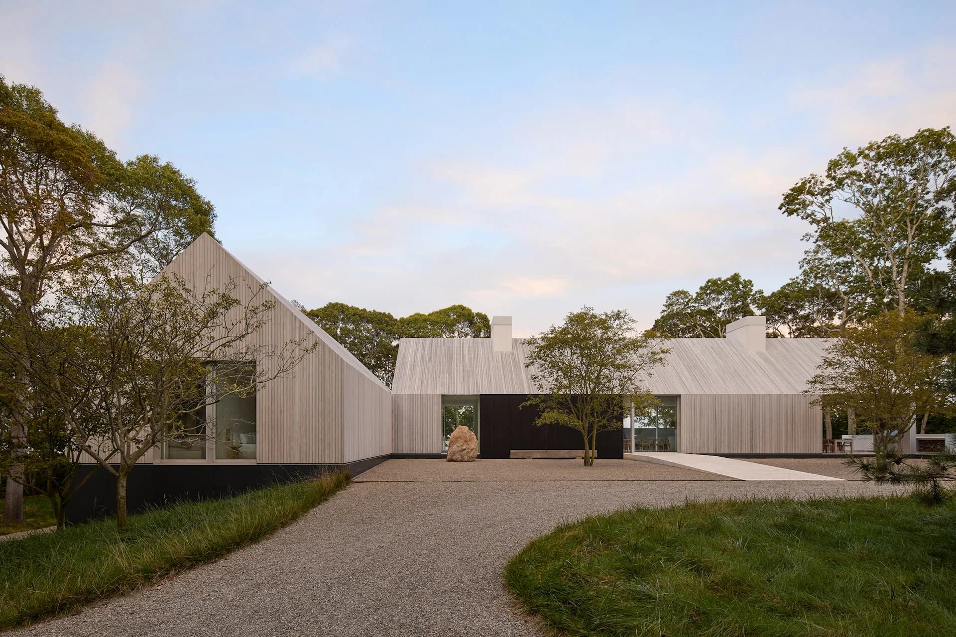 Modern residential pavilion with white corrugated metal cladding and angular roof nestled among mature trees, gravel courtyard entry by 1100 Architect.