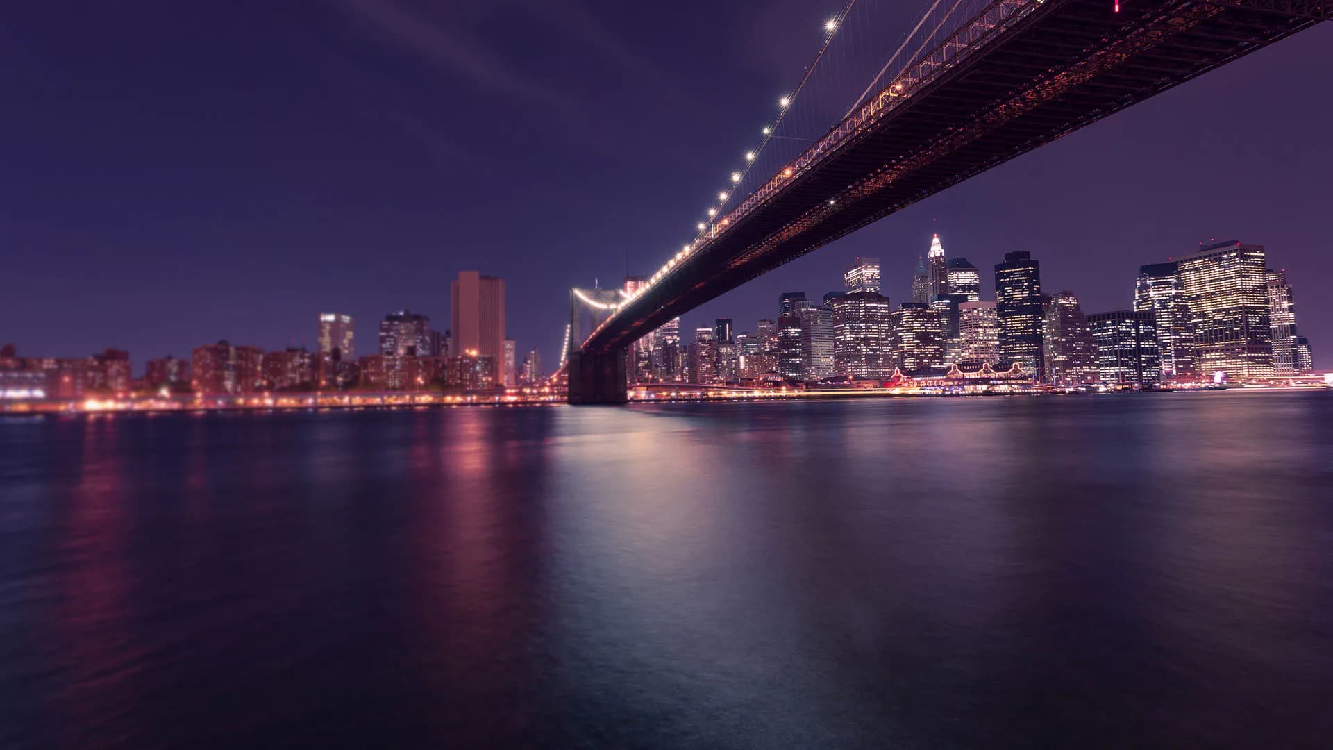 Manhattan skyline and Brooklyn Bridge illuminated at twilight, reflecting on calm East River waters in purple and gold tones by 29 Street Stone Inc.