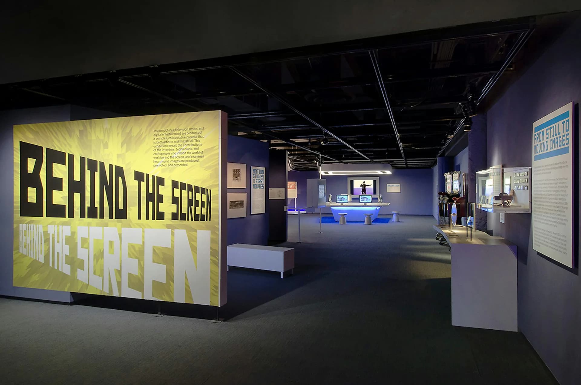 Exhibition interior with dark walls, blue accent lighting, and large yellow signage reading 'BEHIND THE SCREEN' designed by AC Hocek Architecture LLC - (ACHA).