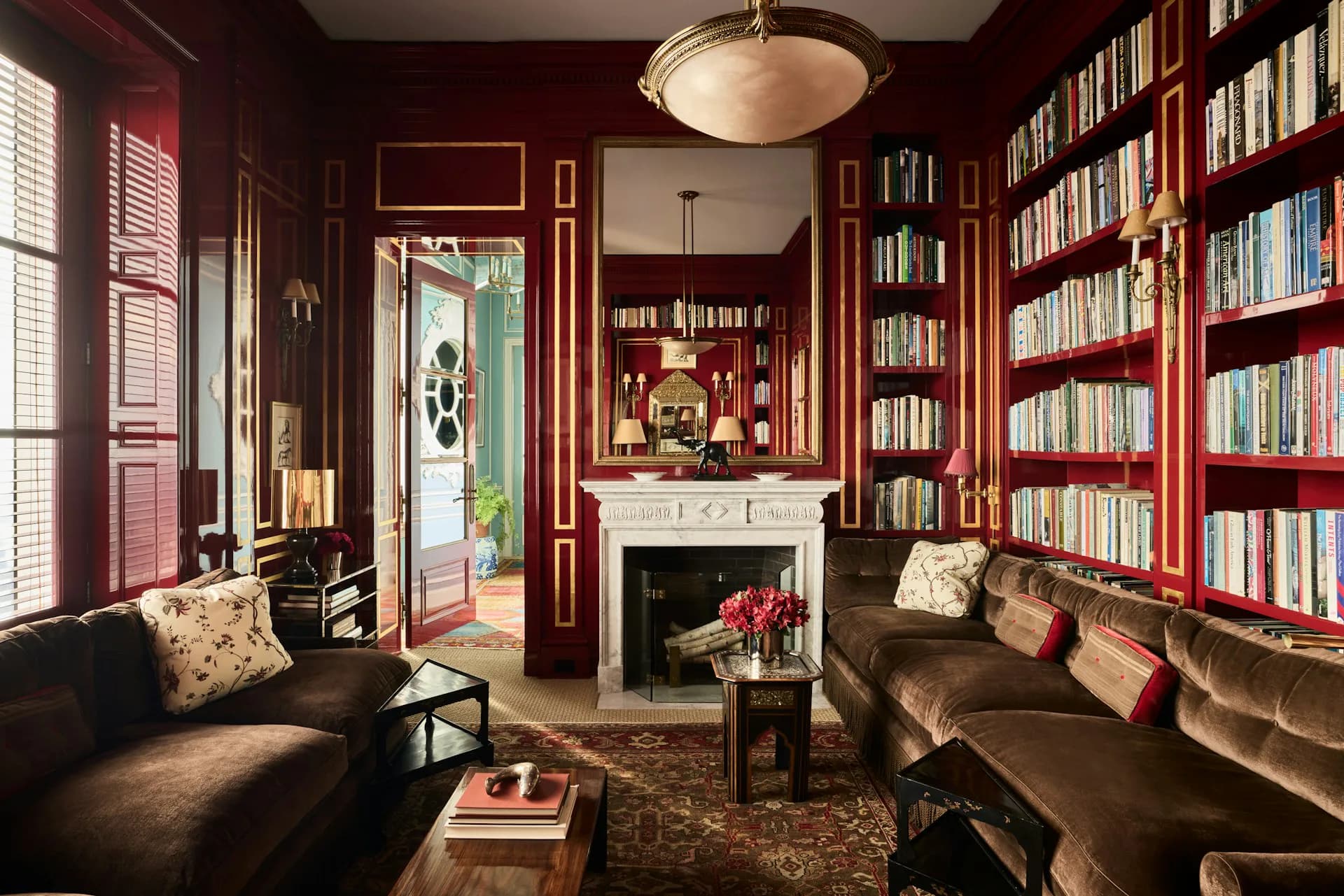 A luxurious home library with deep red walls, floor-to-ceiling bookshelves, dark leather seating, a fireplace, and gold architectural details by Acheson Doyle Partners Architects, P.C.