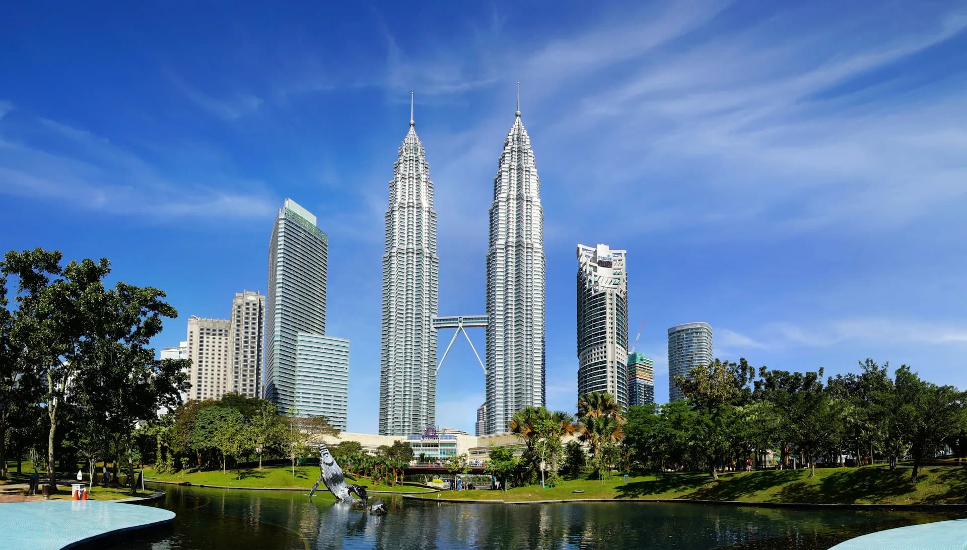 Petronas Twin Towers in Kuala Lumpur rise above a landscaped park with reflective water features and mature trees under blue sky, by Adamson Associates Architects.