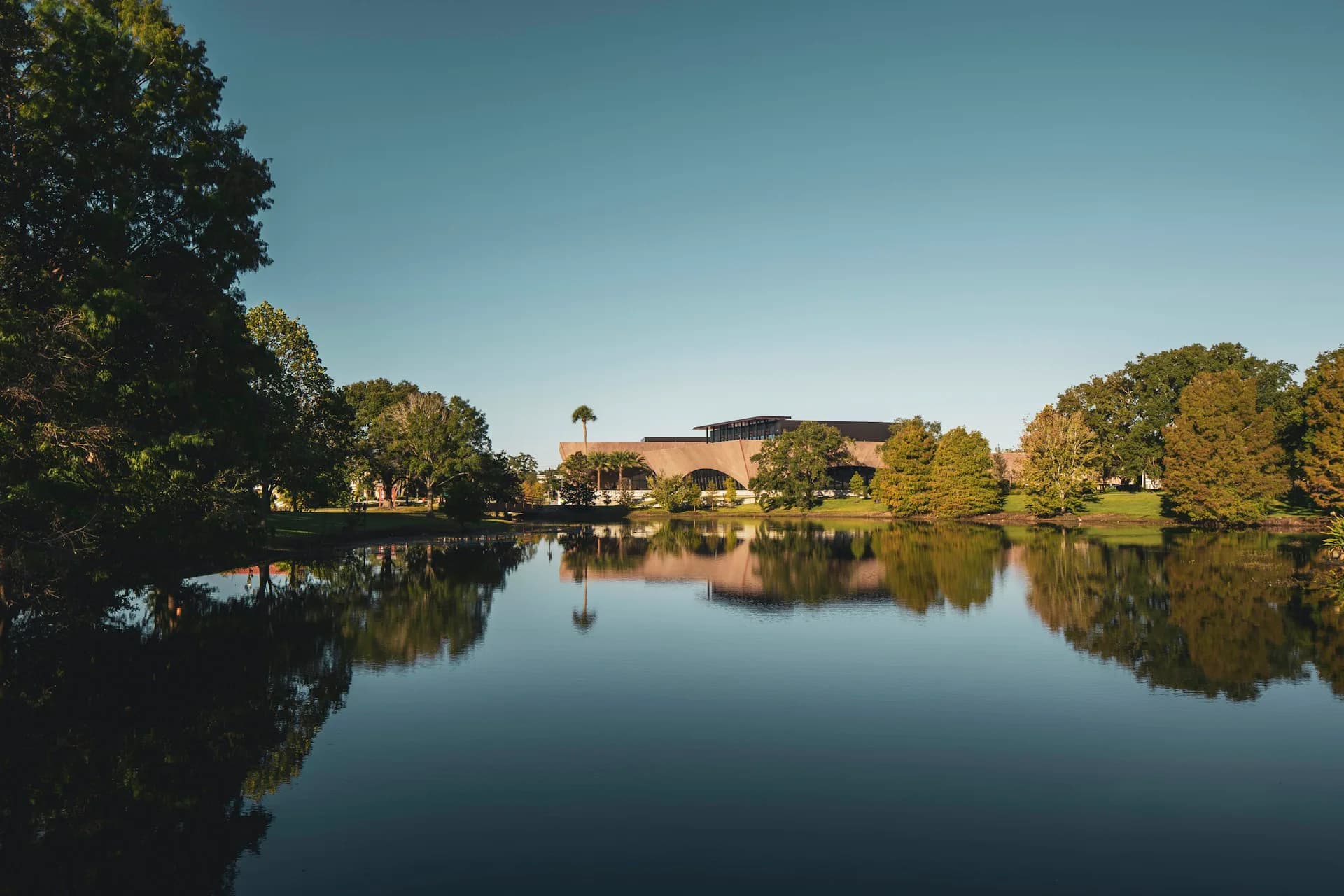Serene riverside landscape with modernist building nestled among mature trees reflected in still water, by Adjaye Associates.