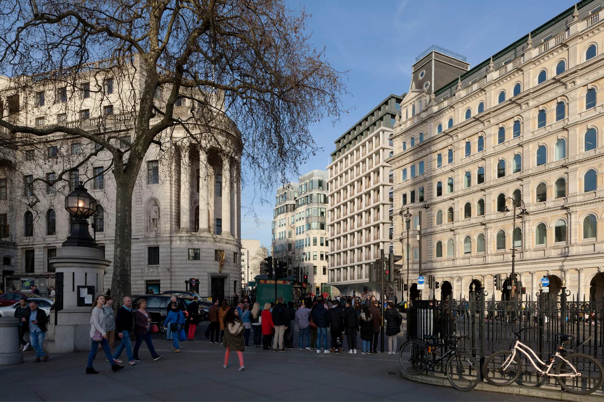 Bustling public plaza with classical stone buildings, bare trees, and crowds of pedestrians in daylight by Adjaye Associates.