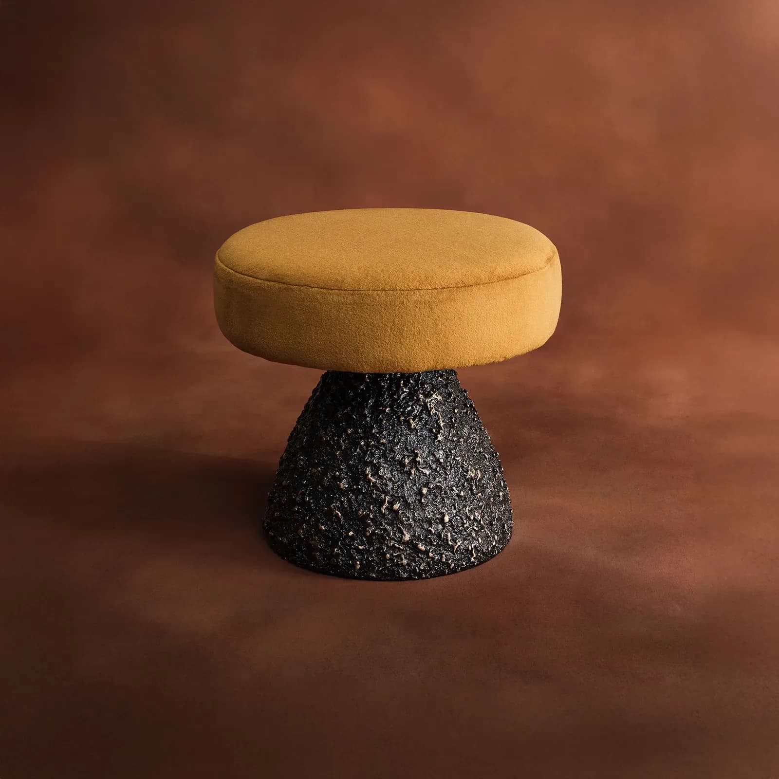 A sculptural stool with a mustard-yellow upholstered circular seat and textured black base against a warm brown backdrop by Aguirre Design.