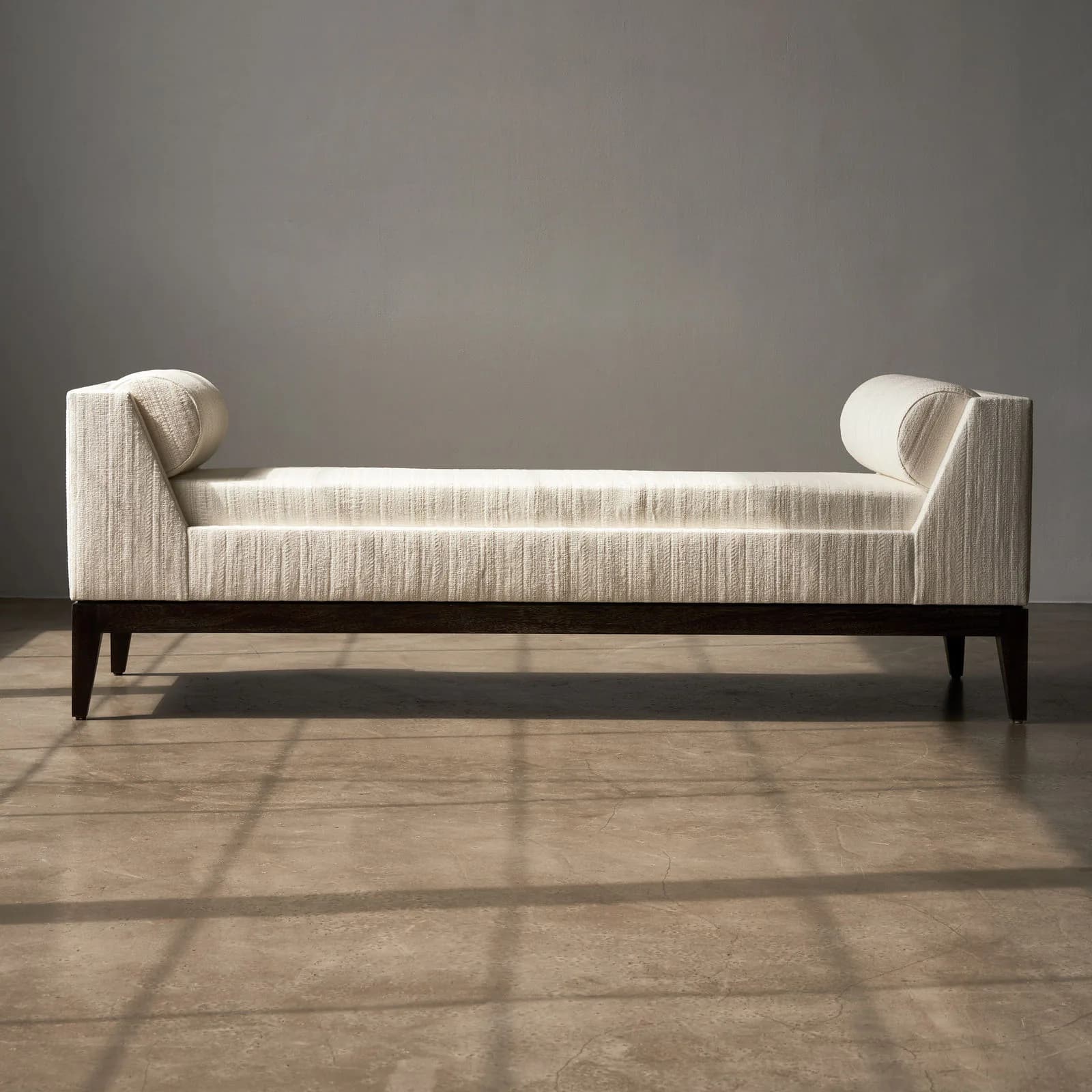 Modern upholstered daybed with cylindrical bolster pillows and dark wooden legs in a minimalist interior space by Aguirre Design.